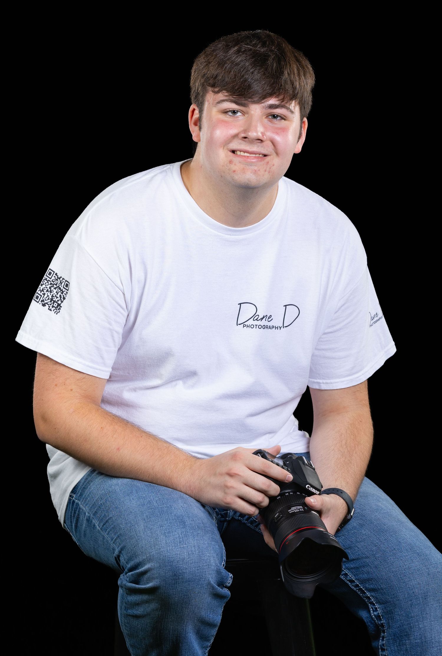 Young photographer smiling in white Dane D Photography t-shirt, holding a camera, seated against black background.