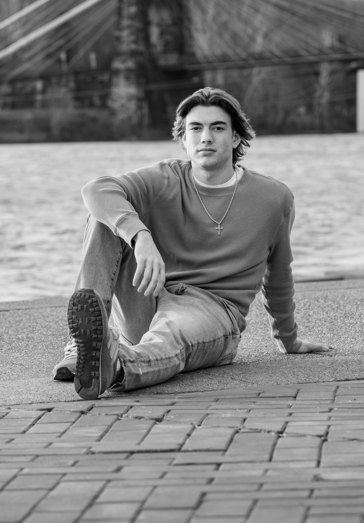 Black and white senior portrait of young man sitting on brick pavement by a river.