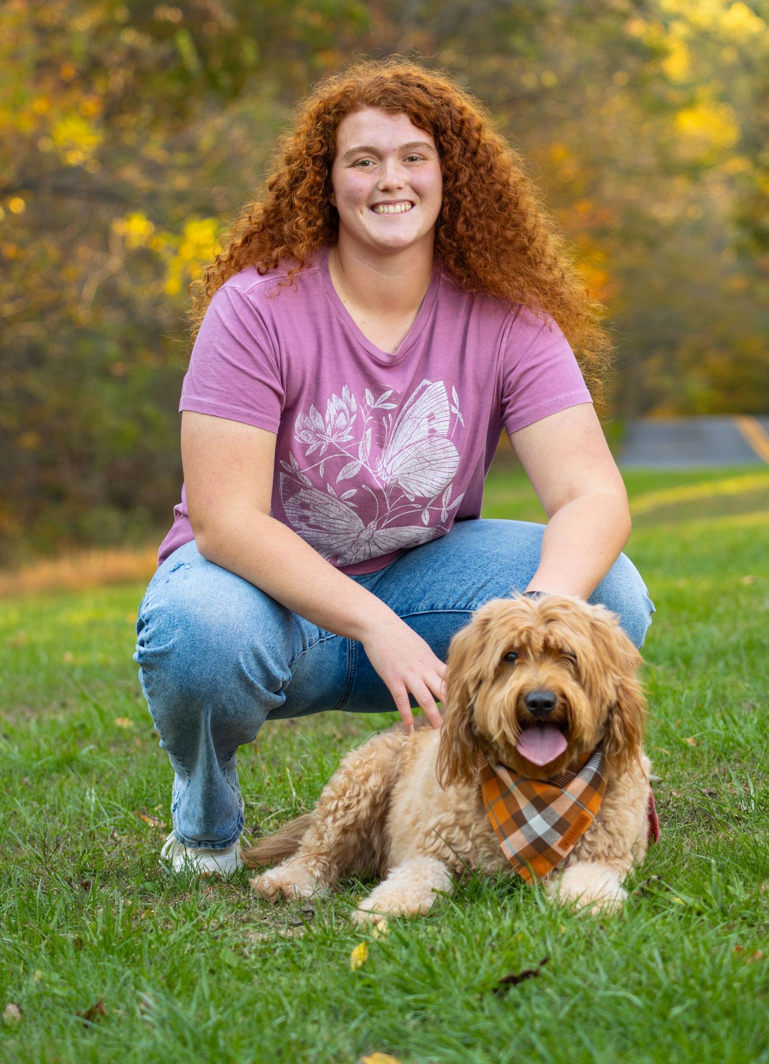 Smiling girl with curly red hair crouches on grass with a fluffy golden doodle dog in autumn park.