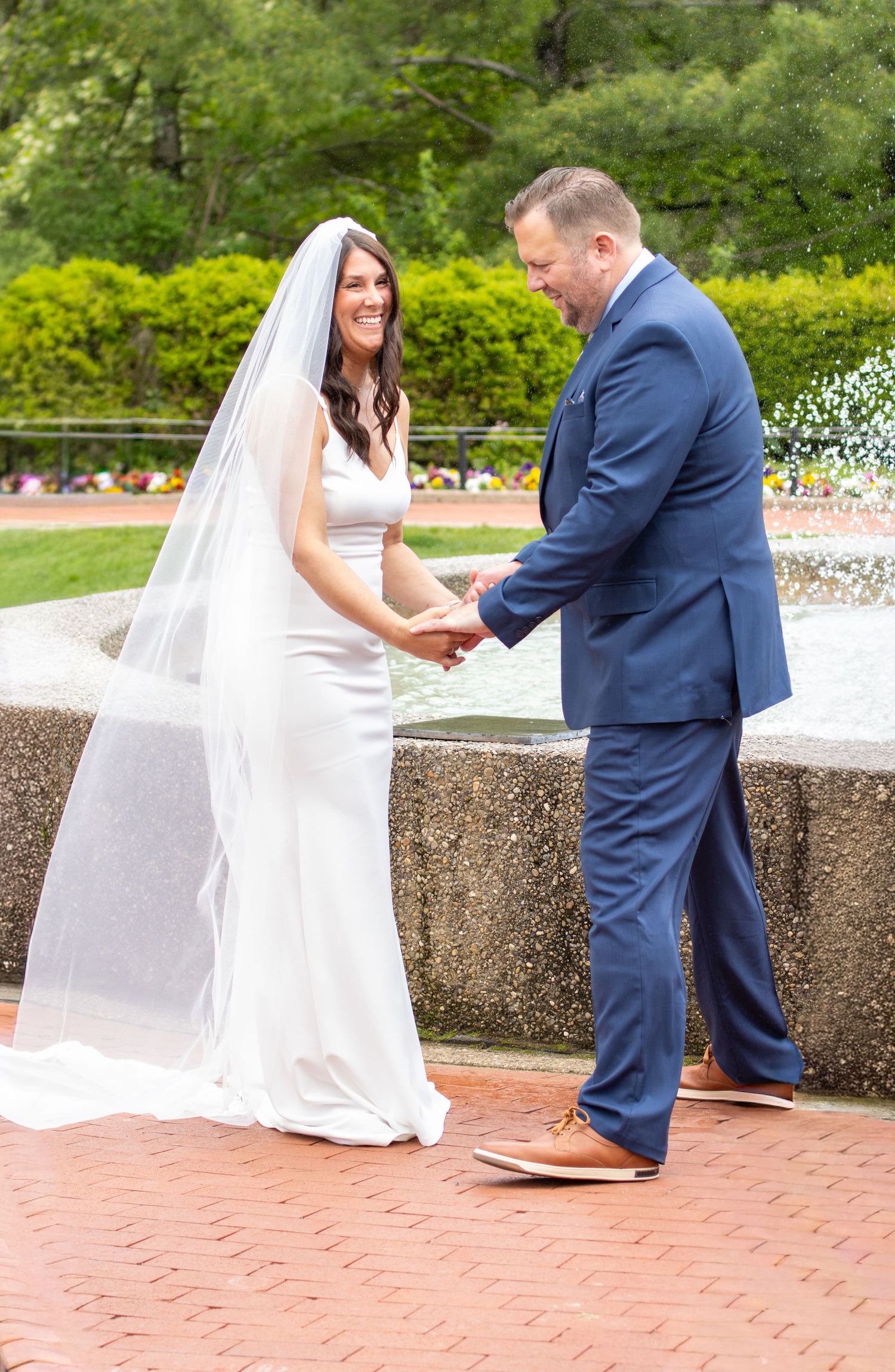 Bride in white gown and veil holds hands with groom in blue suit by fountain.