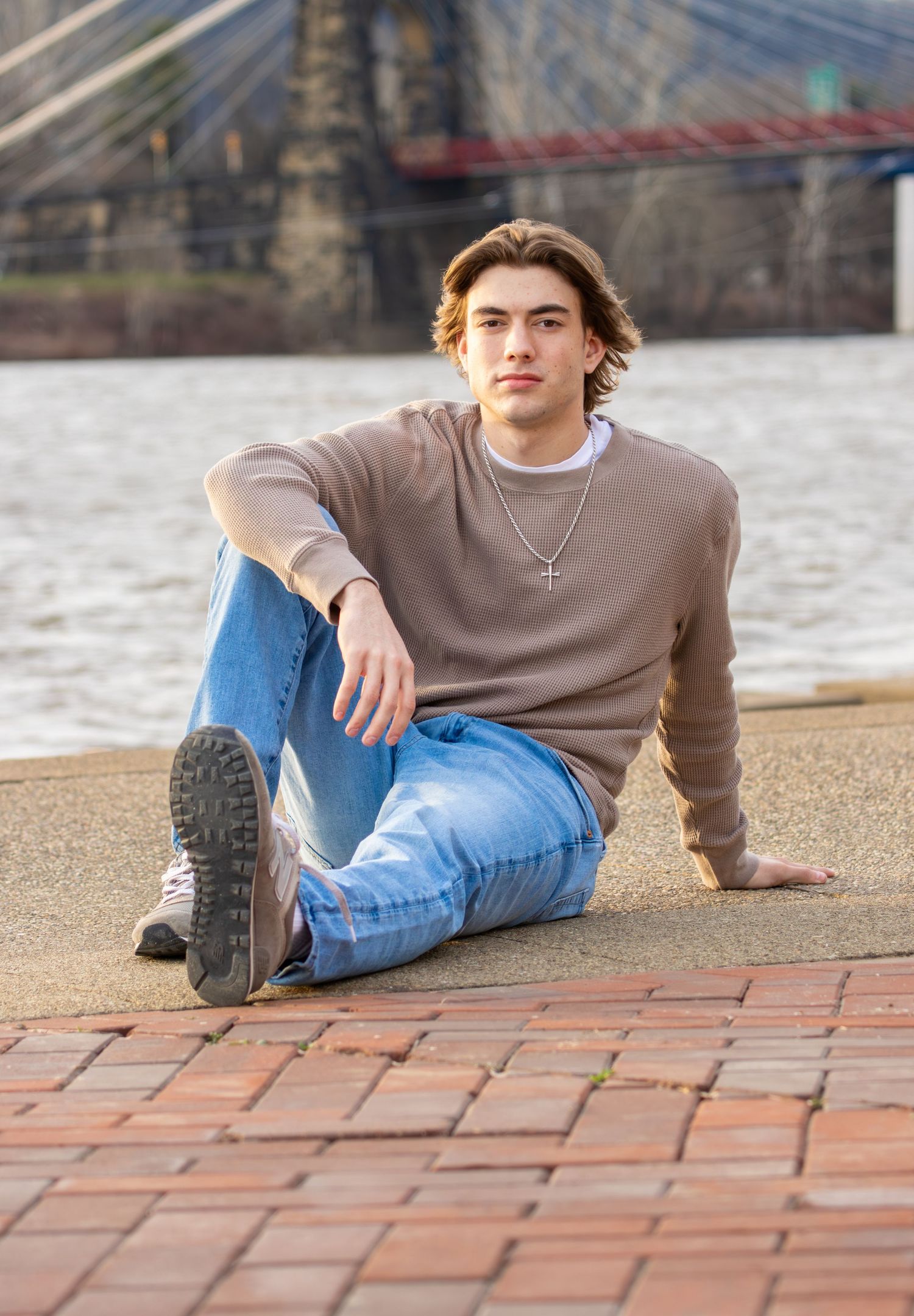 Teen boy in beige sweater and jeans sitting on brick pavement by a river for senior portraits.