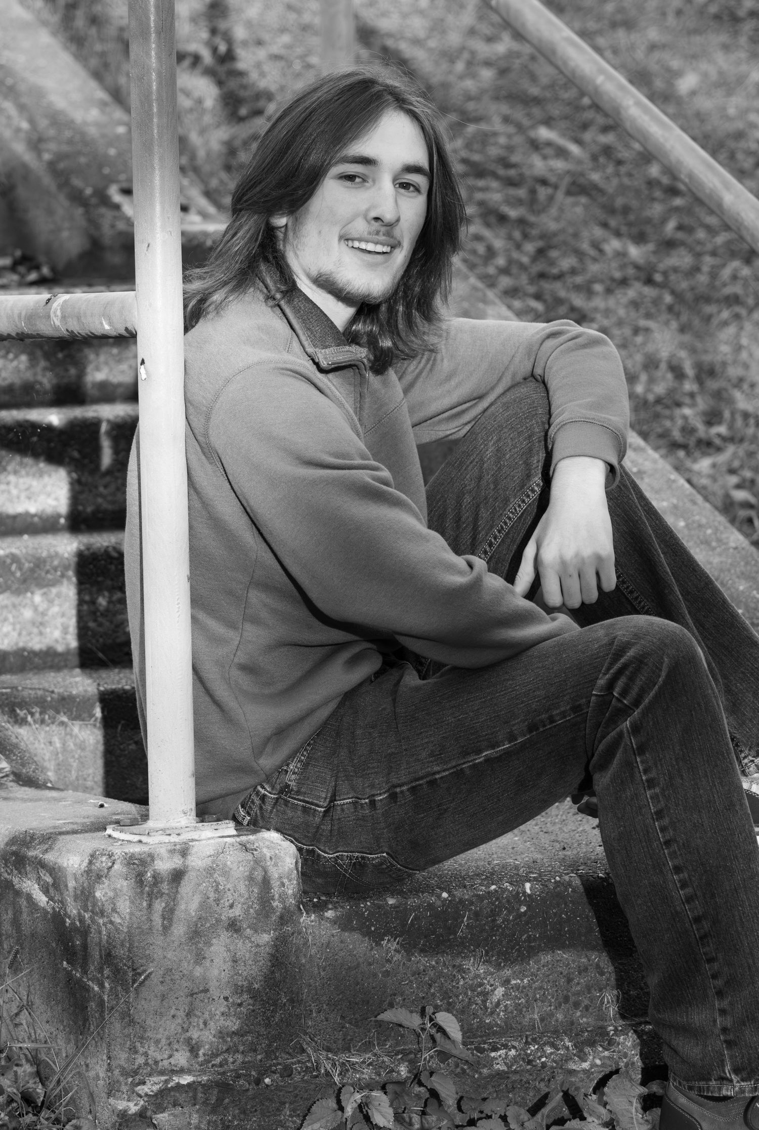 Black and white portrait of a smiling young man with long hair sitting on stone steps.