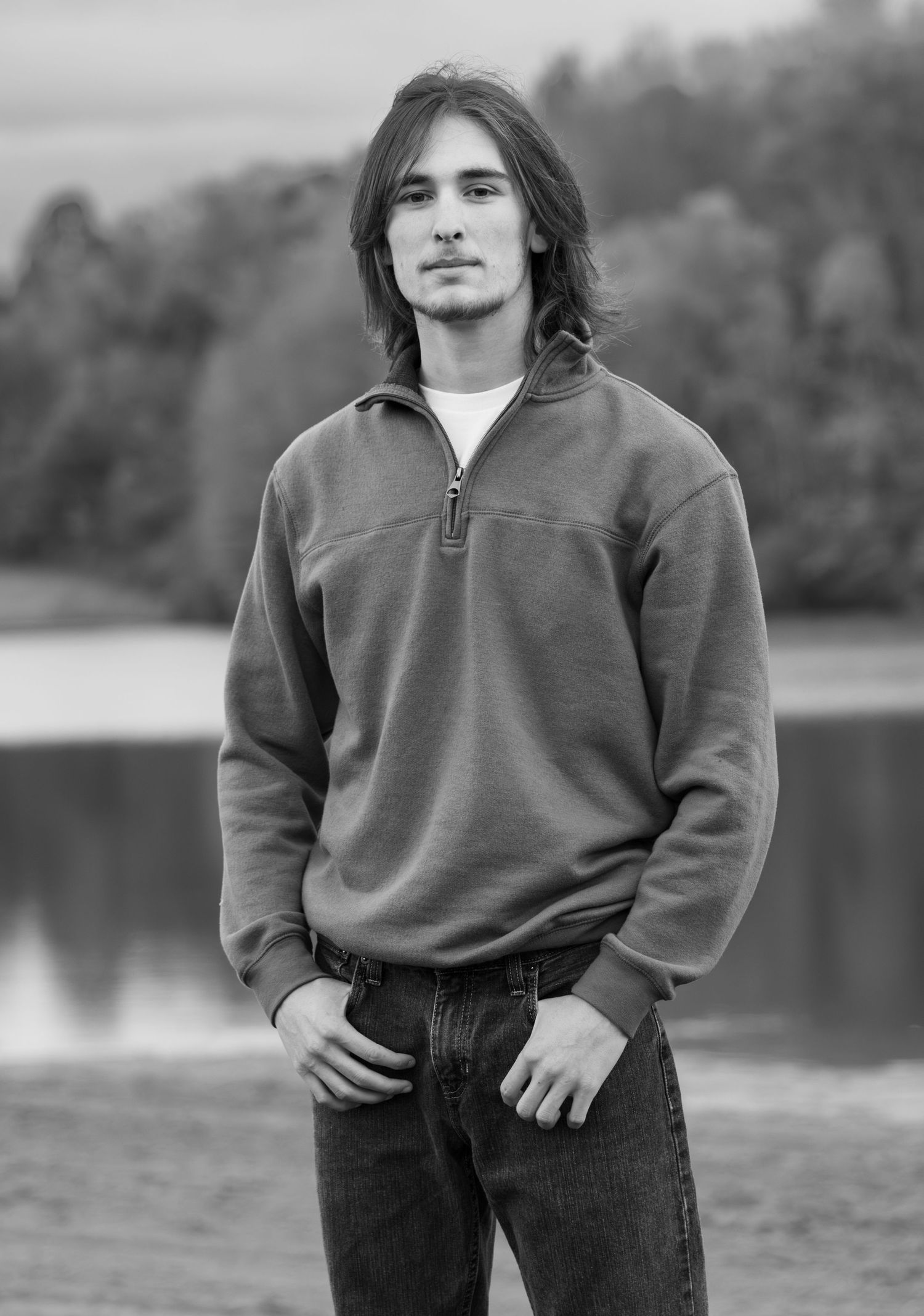 Young man with long hair poses in black and white near a lake wearing a quarter-zip sweatshirt and jeans.