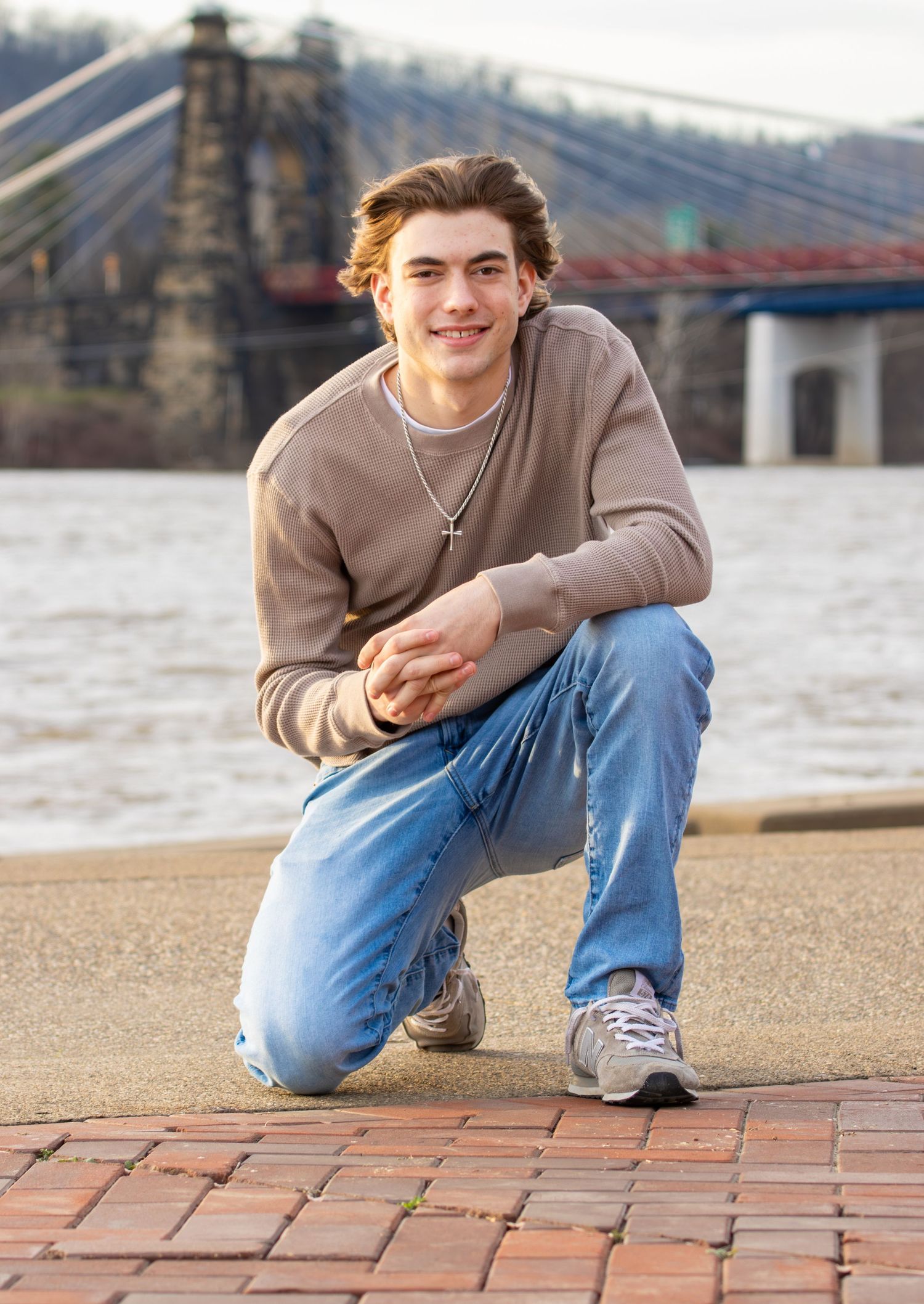 Smiling teen boy in beige sweater kneeling on brick pavement with river bridge backdrop.