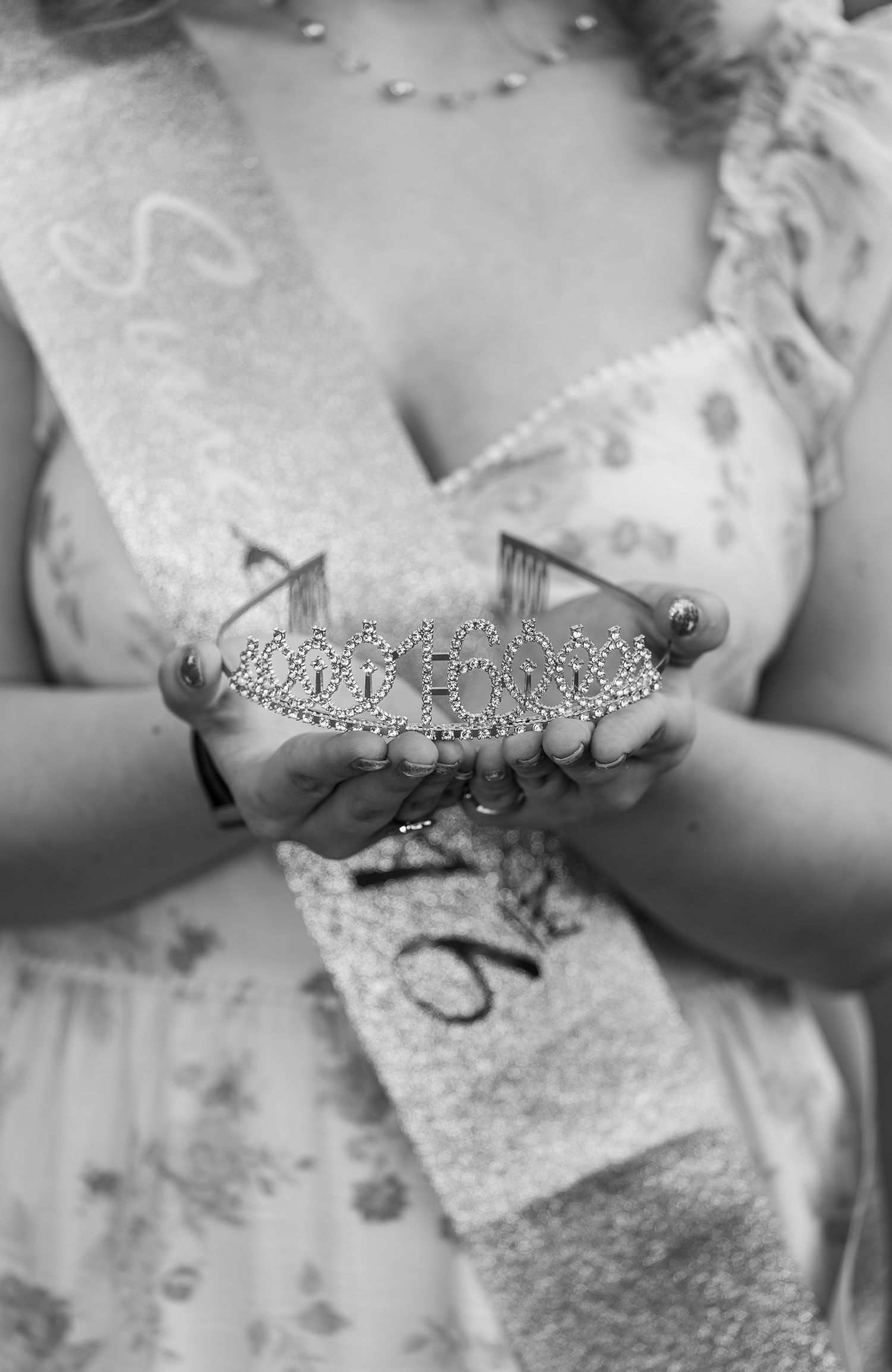 Black and white close-up of hands holding a rhinestone tiara with a Sweet 16 sash.