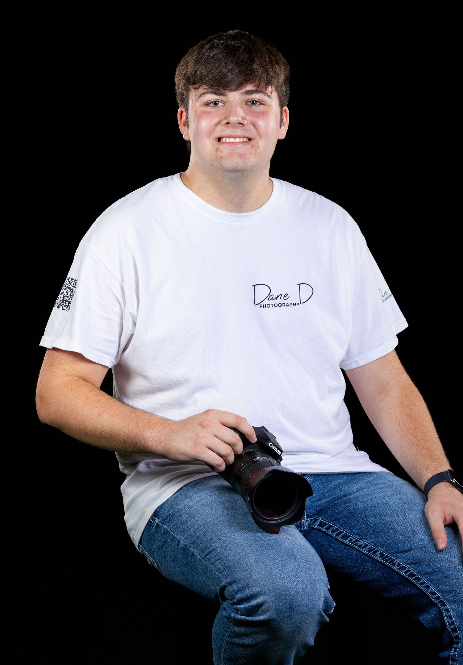 Young photographer smiling in white Dane D Photography t-shirt, holding camera, seated against black background.