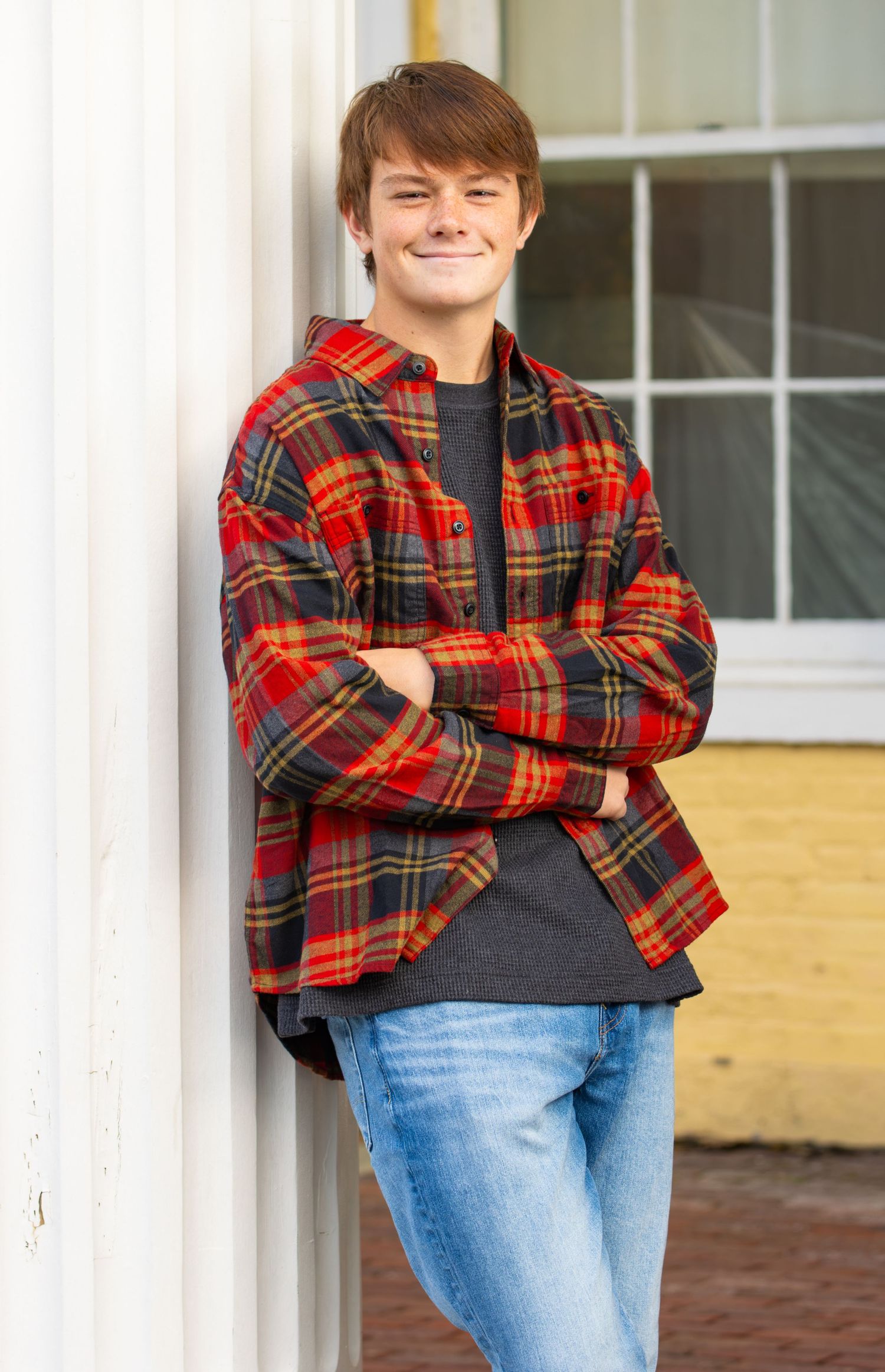 Smiling teen boy in red plaid flannel shirt leaning against white pillar outdoors.
