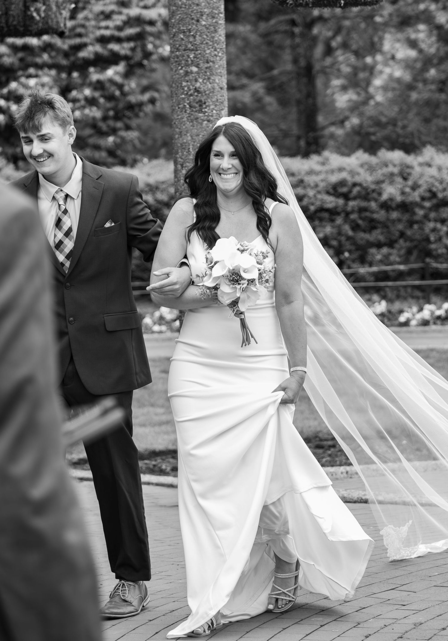 Smiling bride in white gown and veil walking with escort in black and white photo.