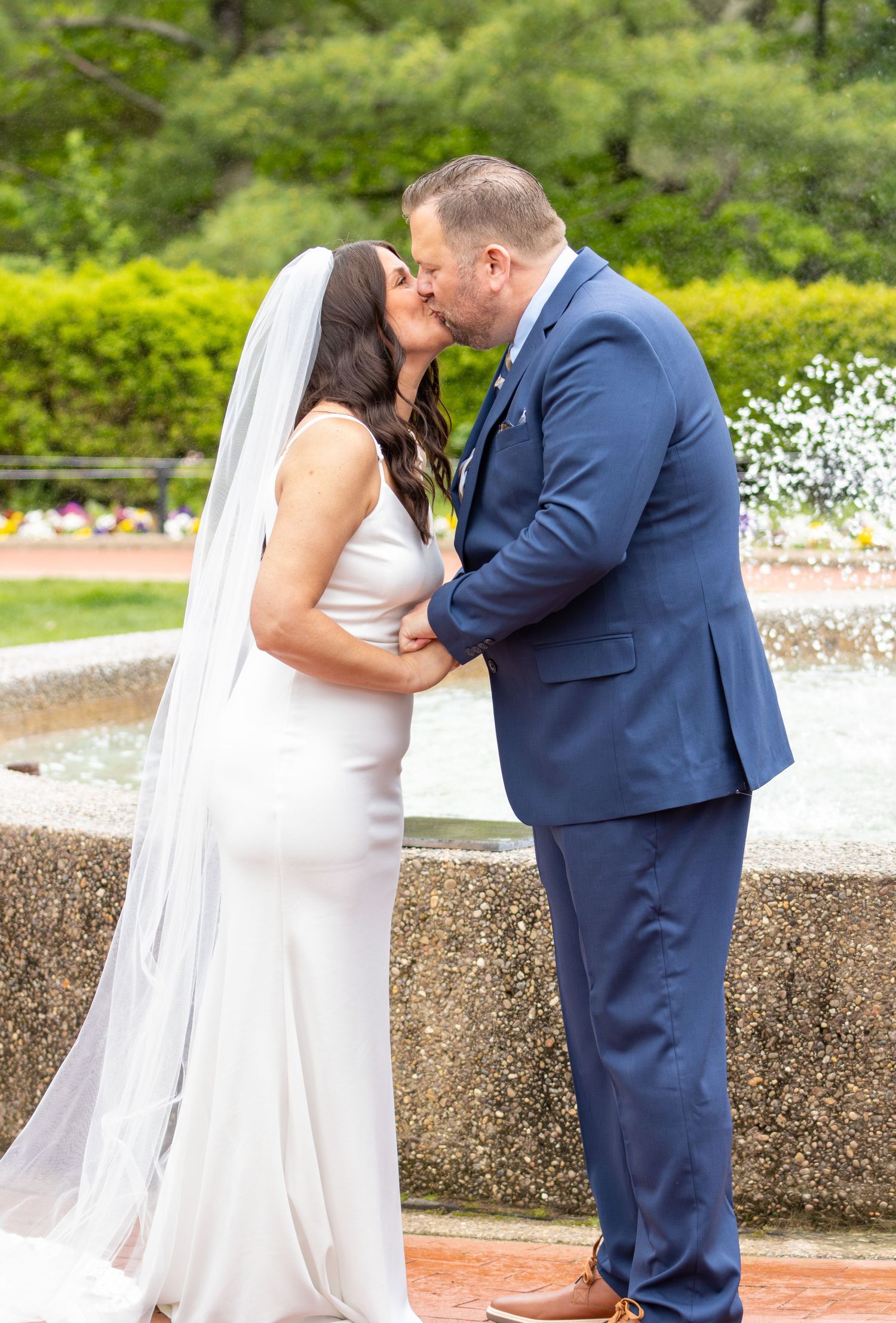 Bride in white fitted gown and groom in blue suit kissing by outdoor fountain.