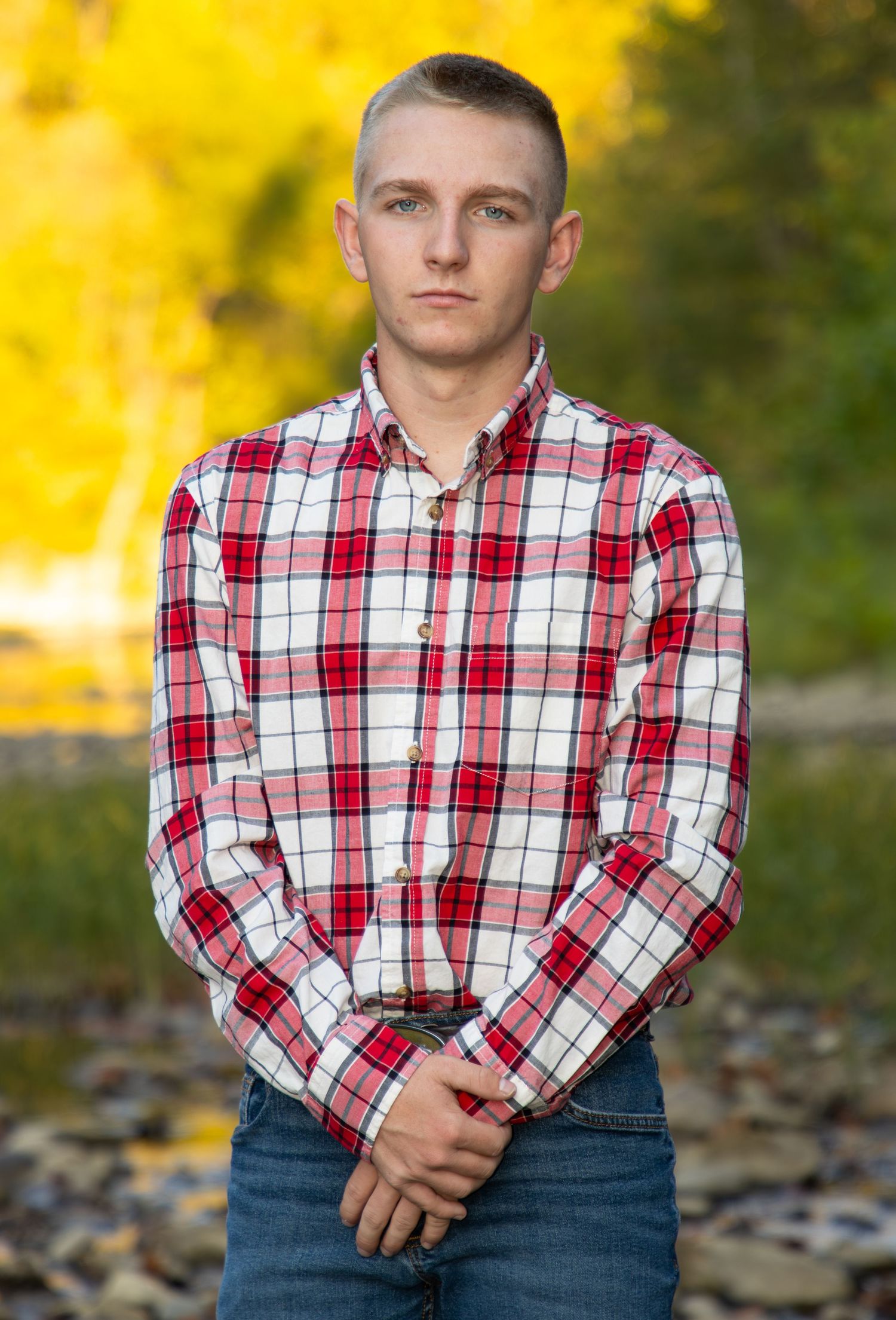 Teen boy in red and white plaid shirt standing outdoors with fall foliage background.