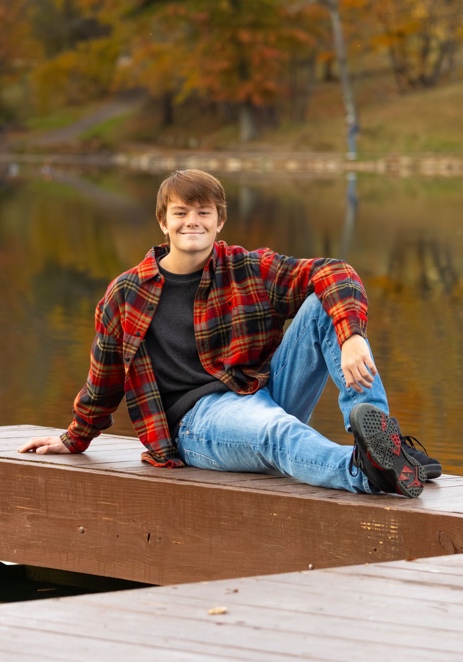Teen boy in red plaid shirt sitting on wooden dock by autumn lake smiling.