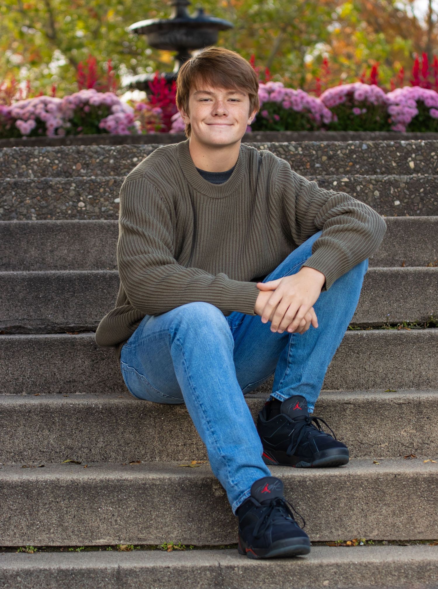 Smiling young man in olive sweater sitting on stone steps with flowers in background.