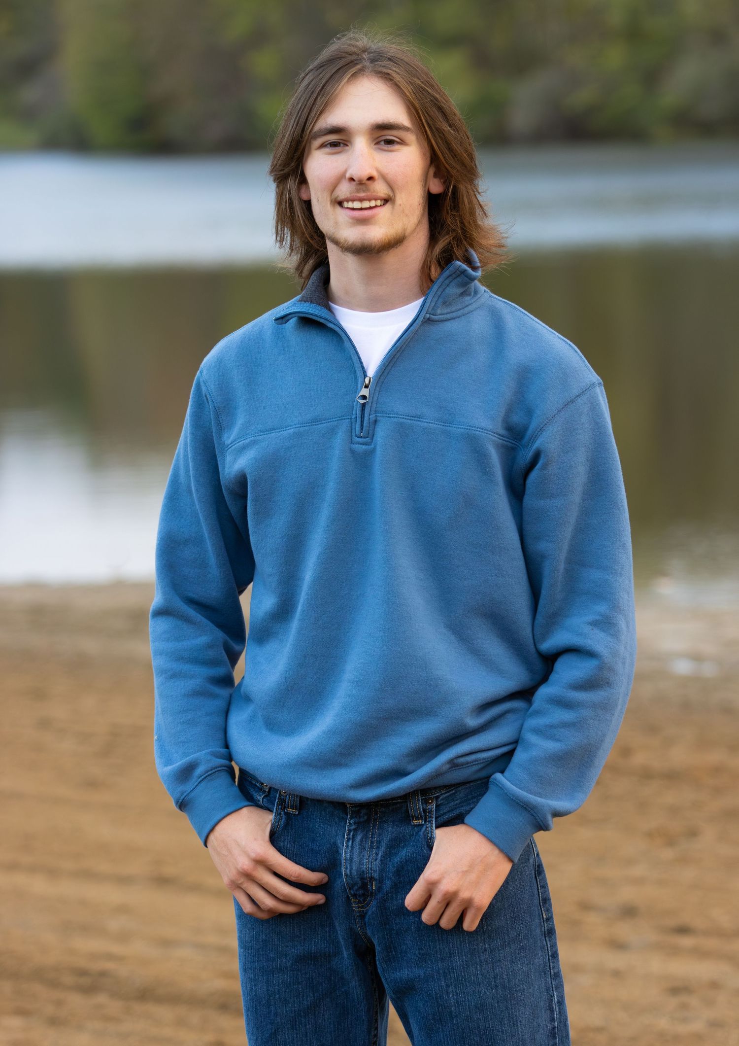 Young man with medium-length hair wearing a blue quarter-zip pullover standing by a lake.