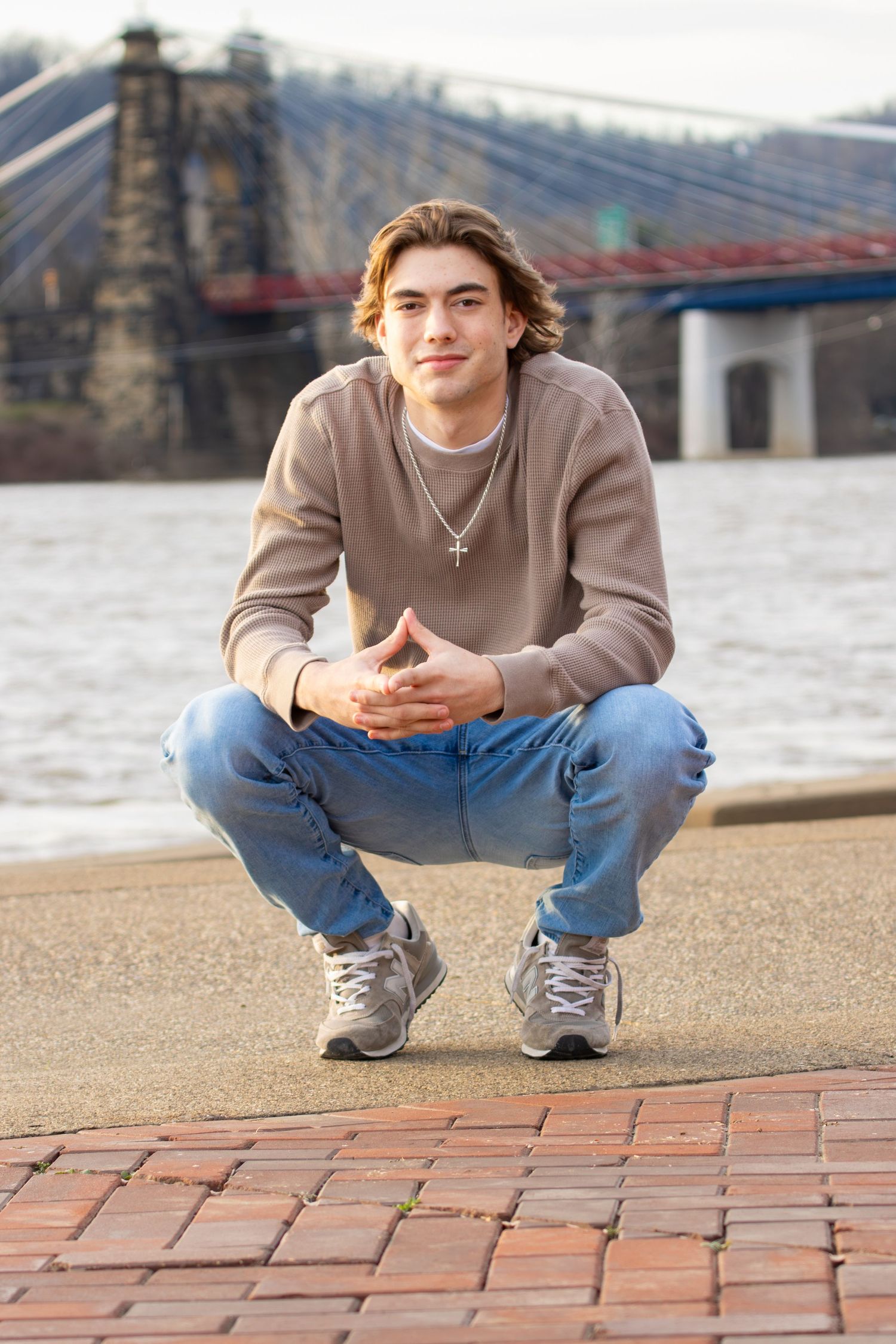 Young man in beige sweater crouching on brick pavement with suspension bridge in background.