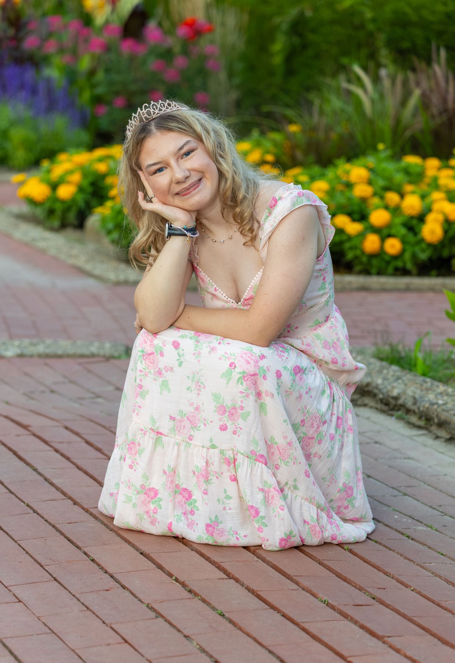 Smiling teenage girl in floral dress and tiara posing on brick path in garden setting.