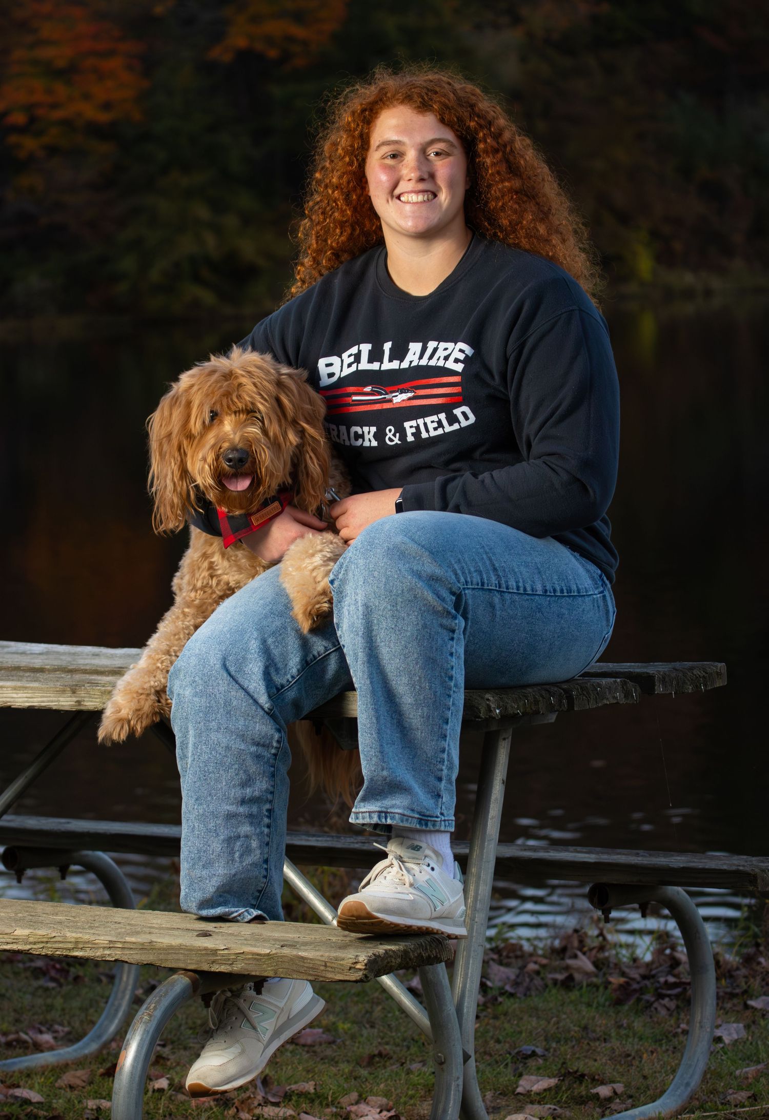 Curly red-haired girl sits on picnic table holding a golden doodle dog wearing a track shirt.