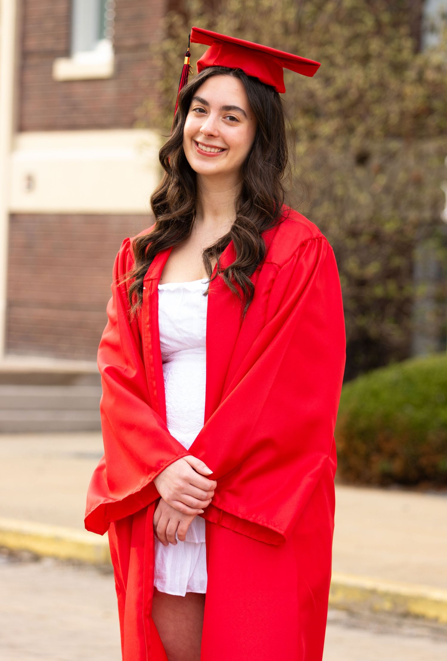 Young woman smiling in red graduation cap and gown over white dress outdoors.