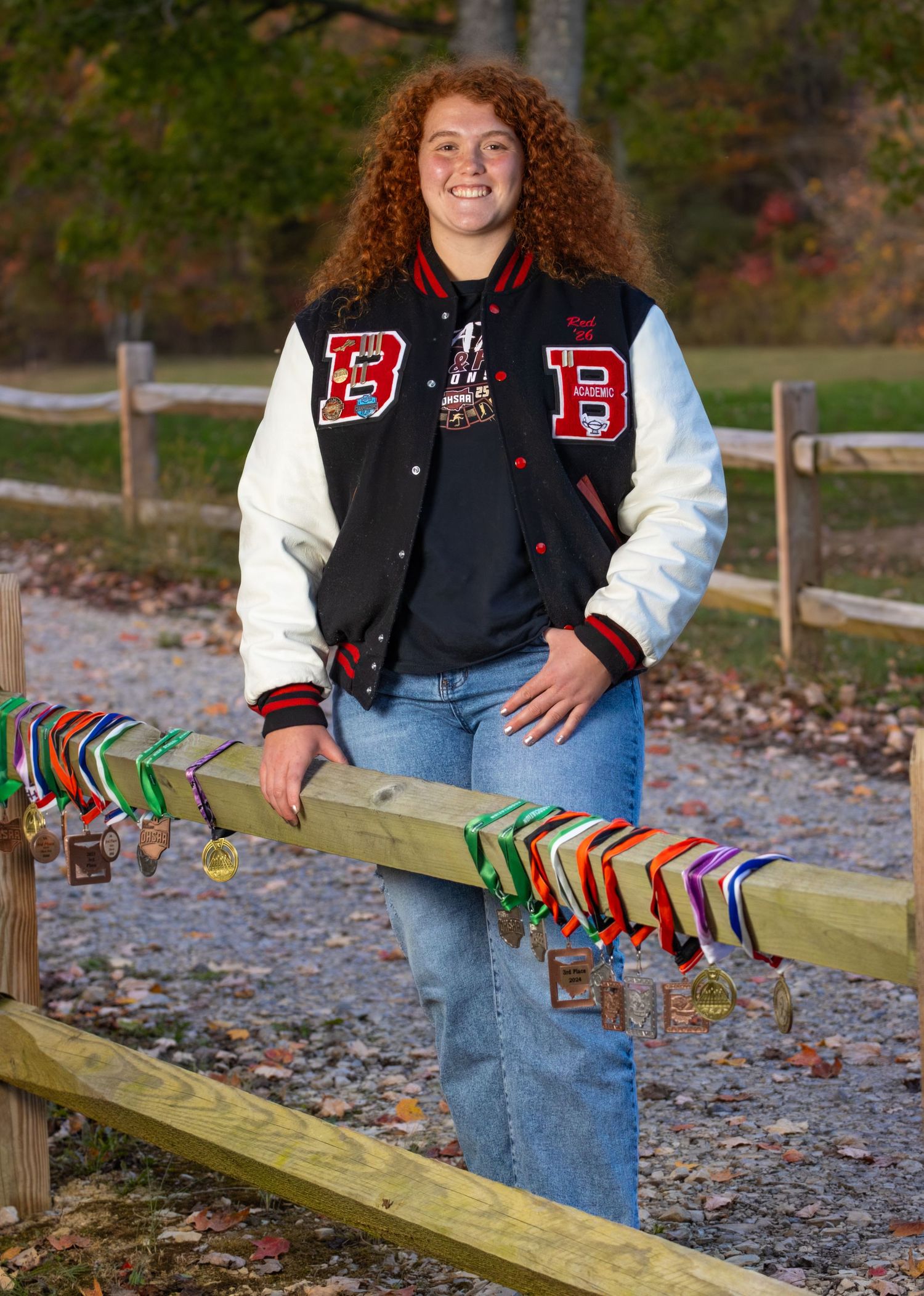 Teen girl with curly red hair wears a varsity letterman jacket displaying medals on a wooden fence.