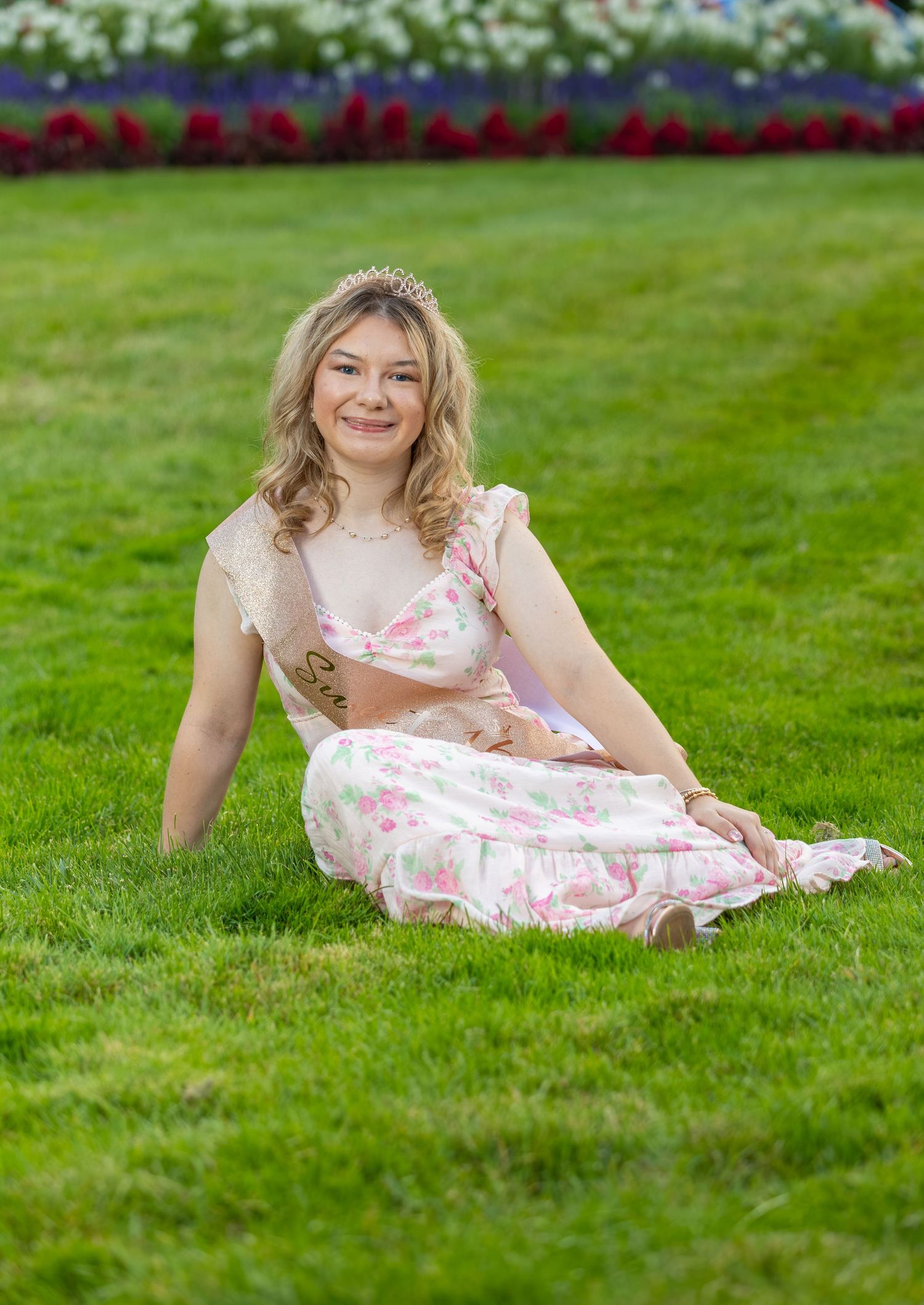Smiling teenage girl in floral dress and tiara sitting on green grass for Sweet 16 portraits.