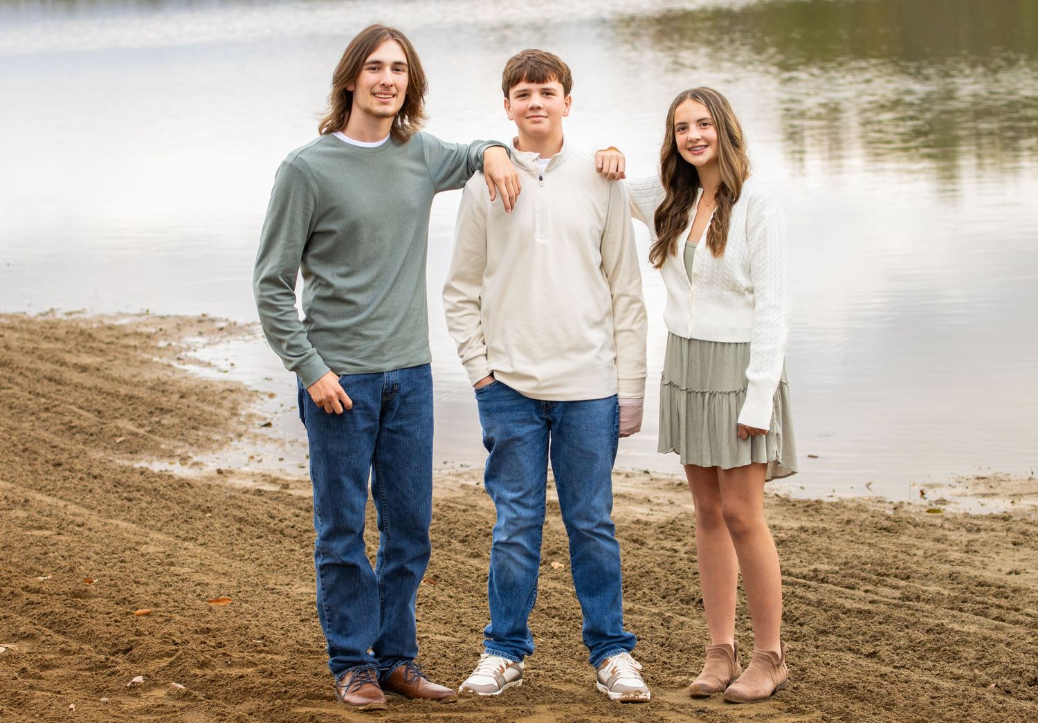 Three siblings standing together on a sandy lakeshore for an outdoor portrait photo.