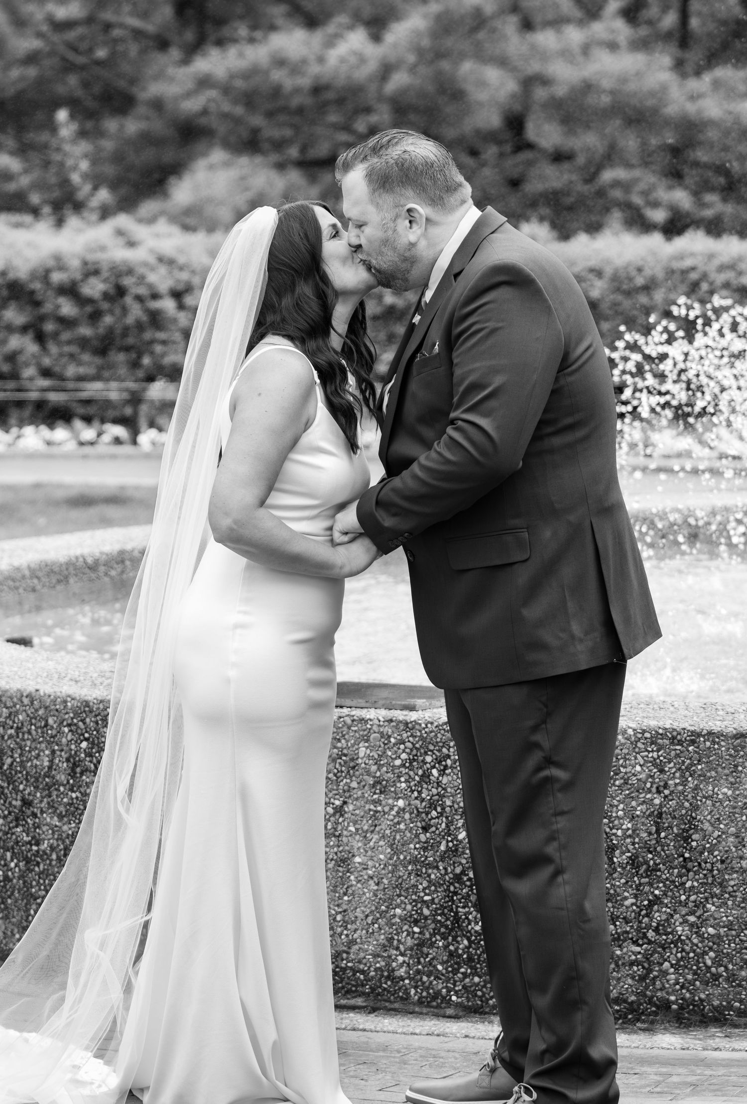 Bride and groom share first kiss in black and white wedding photo by fountain.