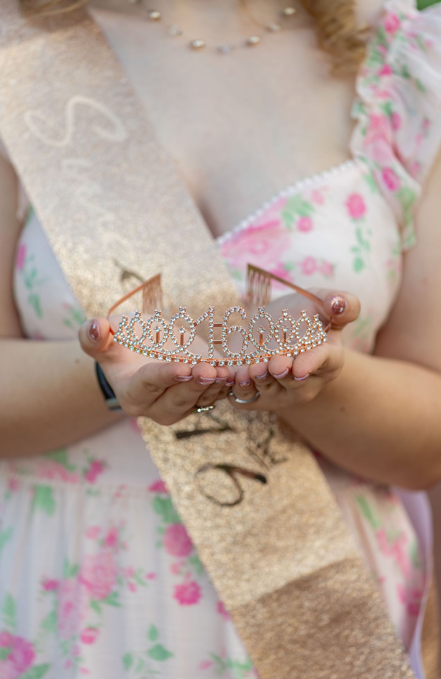 Close-up of hands holding a rhinestone tiara with a gold Sweet 16 sash.