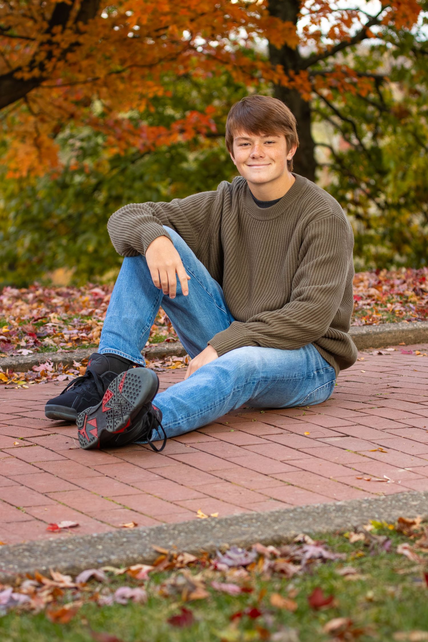 Smiling young man in olive sweater sitting on brick path surrounded by fall leaves.