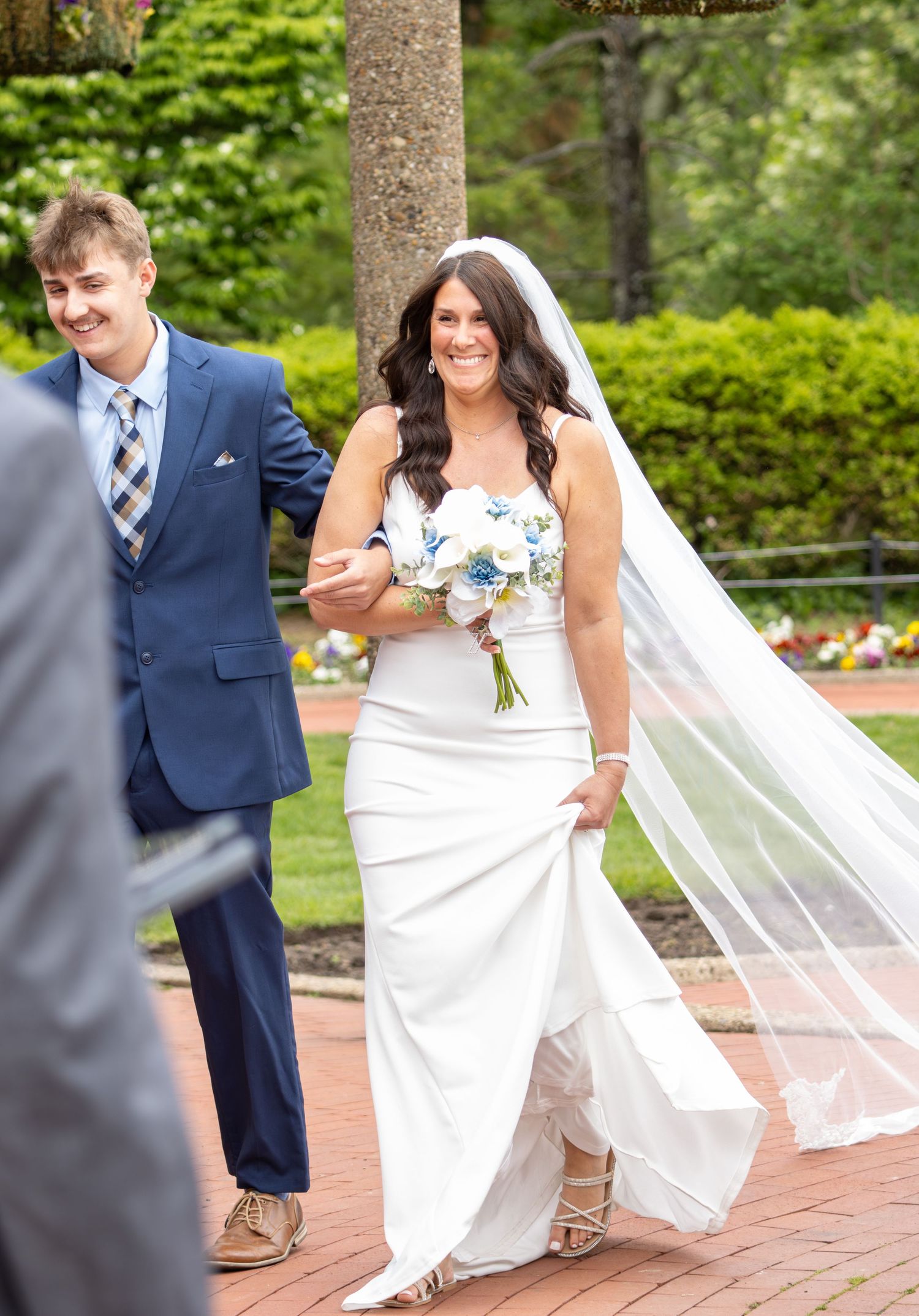Radiant bride in white strapless gown holding bouquet walking with escort outdoors.