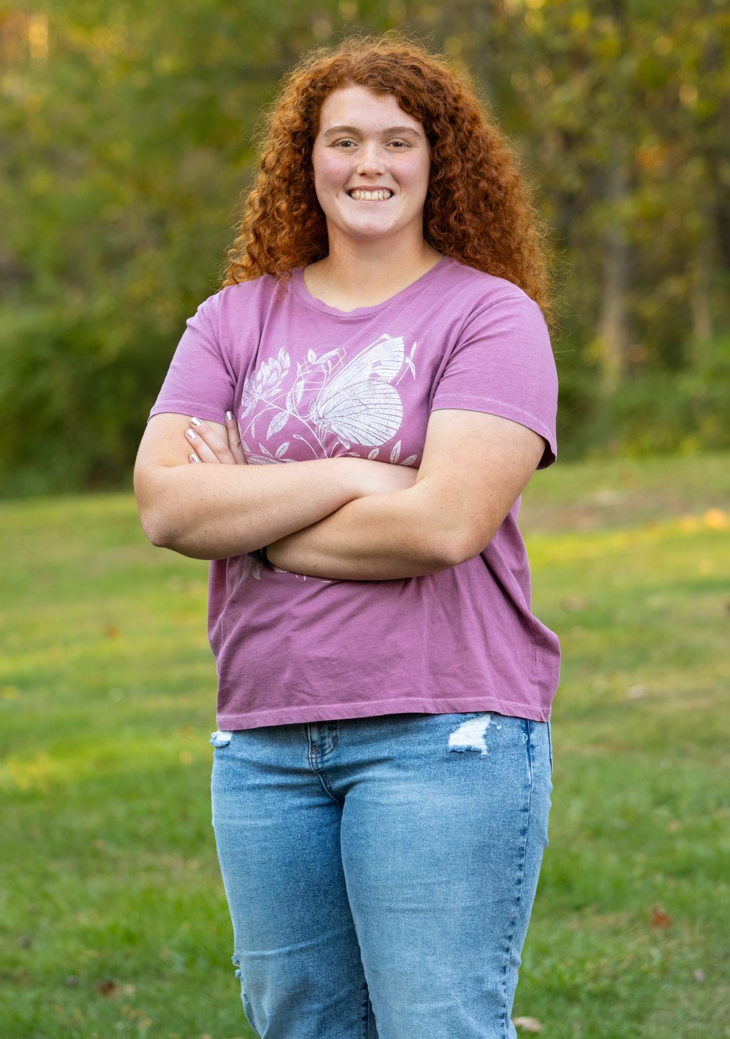 Smiling teenage girl with curly red hair poses outdoors in a purple butterfly t-shirt and jeans.