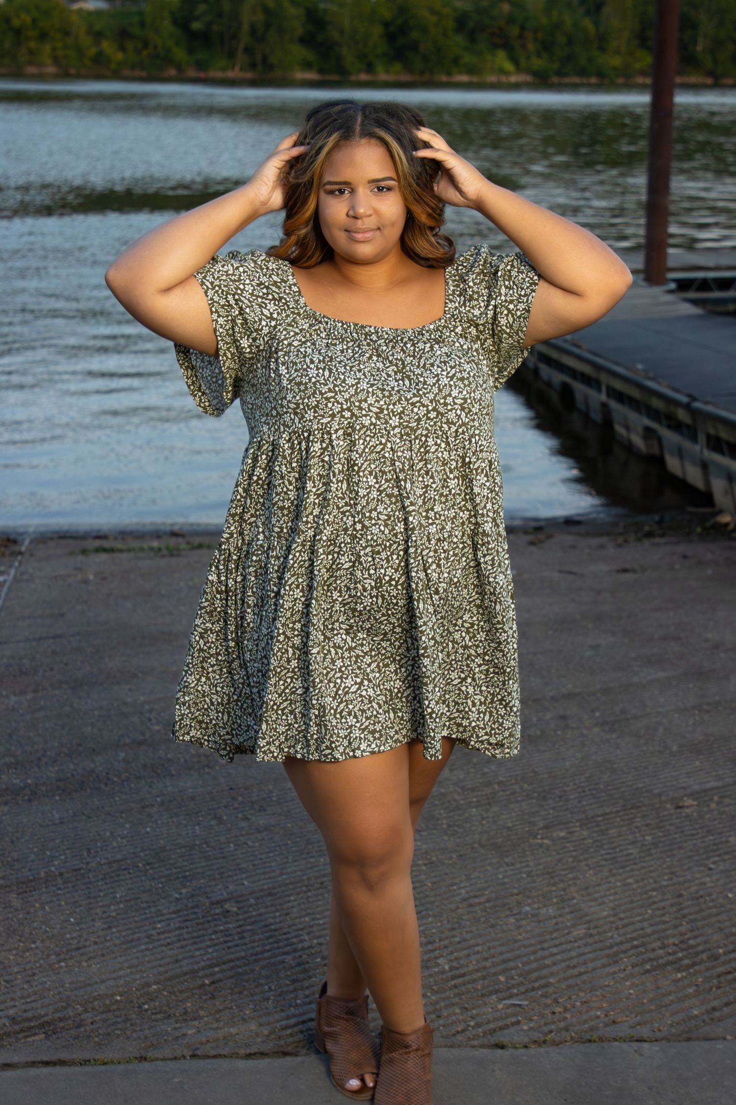Young woman in green floral dress walks near waterfront dock with hands in hair.