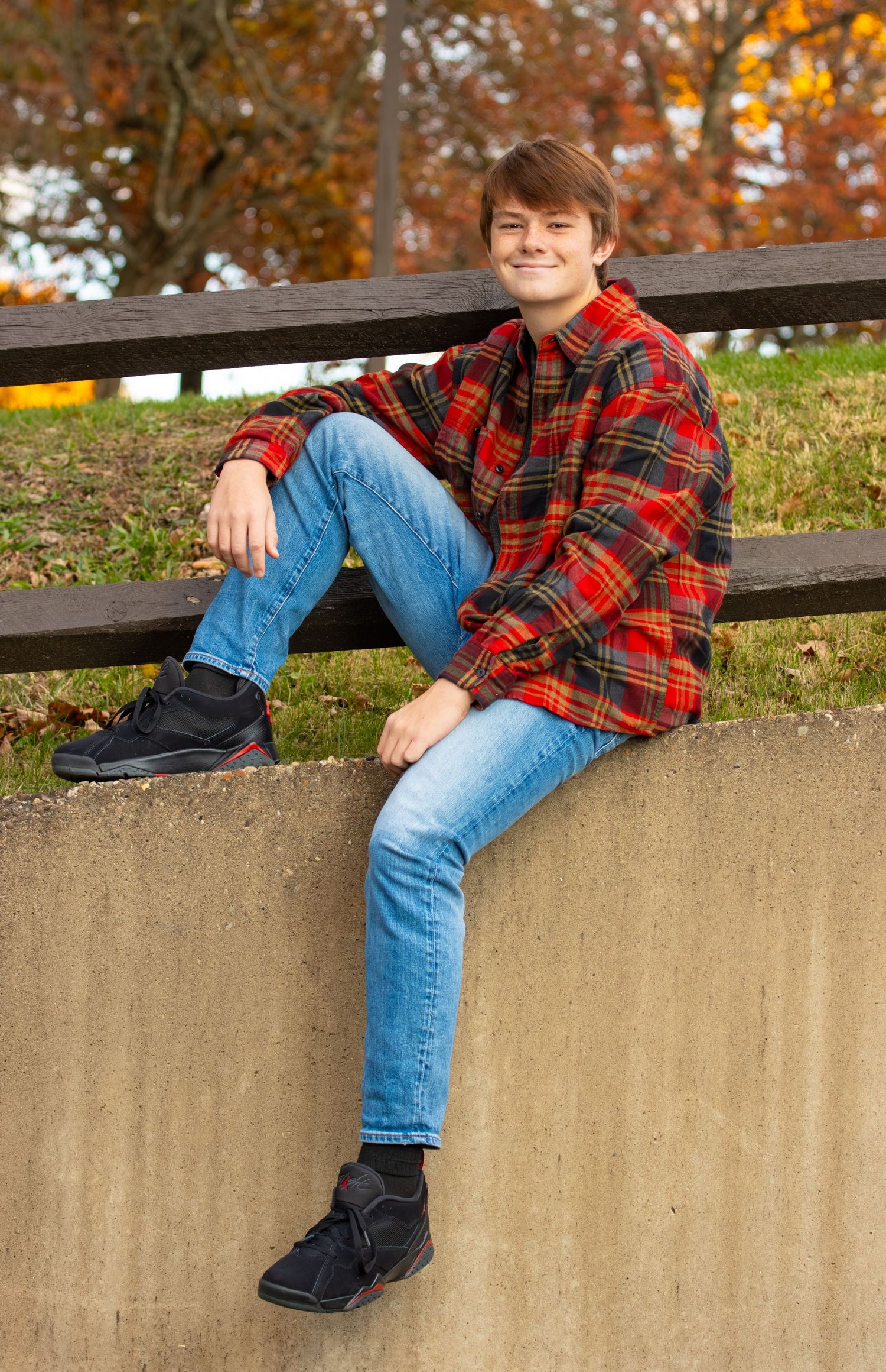 Teen boy in red plaid jacket sitting on concrete wall with autumn trees behind him.