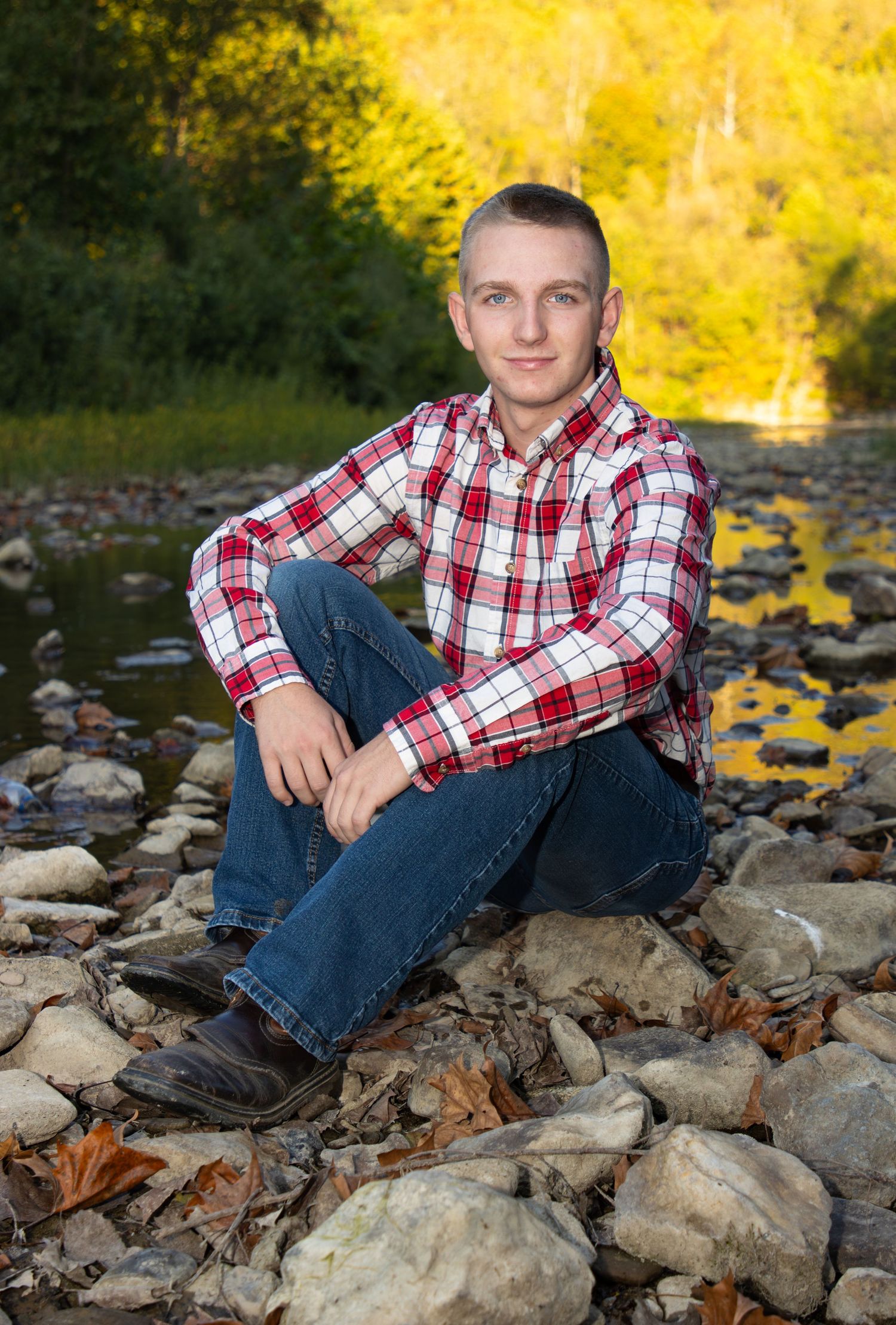 Young man in red plaid shirt sitting on rocks by a stream in autumn.