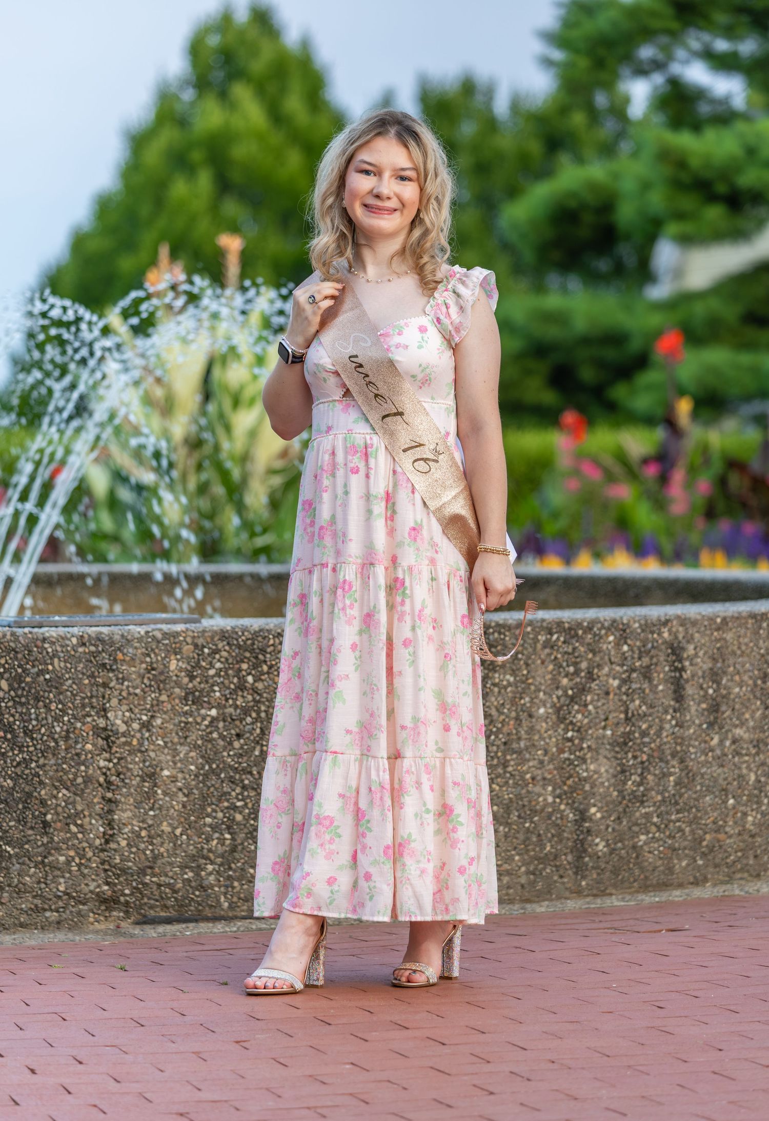 Teen girl in pink floral maxi dress with Sweet 16 sash posing by a fountain outdoors.