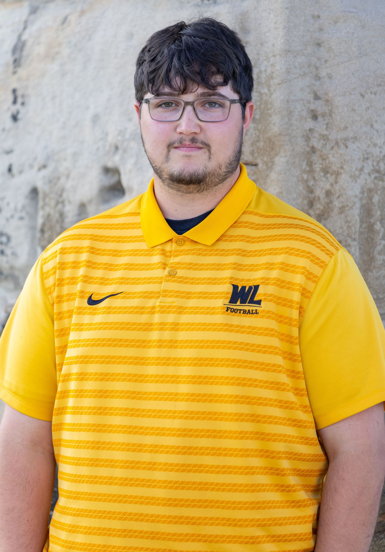 Young man in yellow polo shirt posing against stone wall.