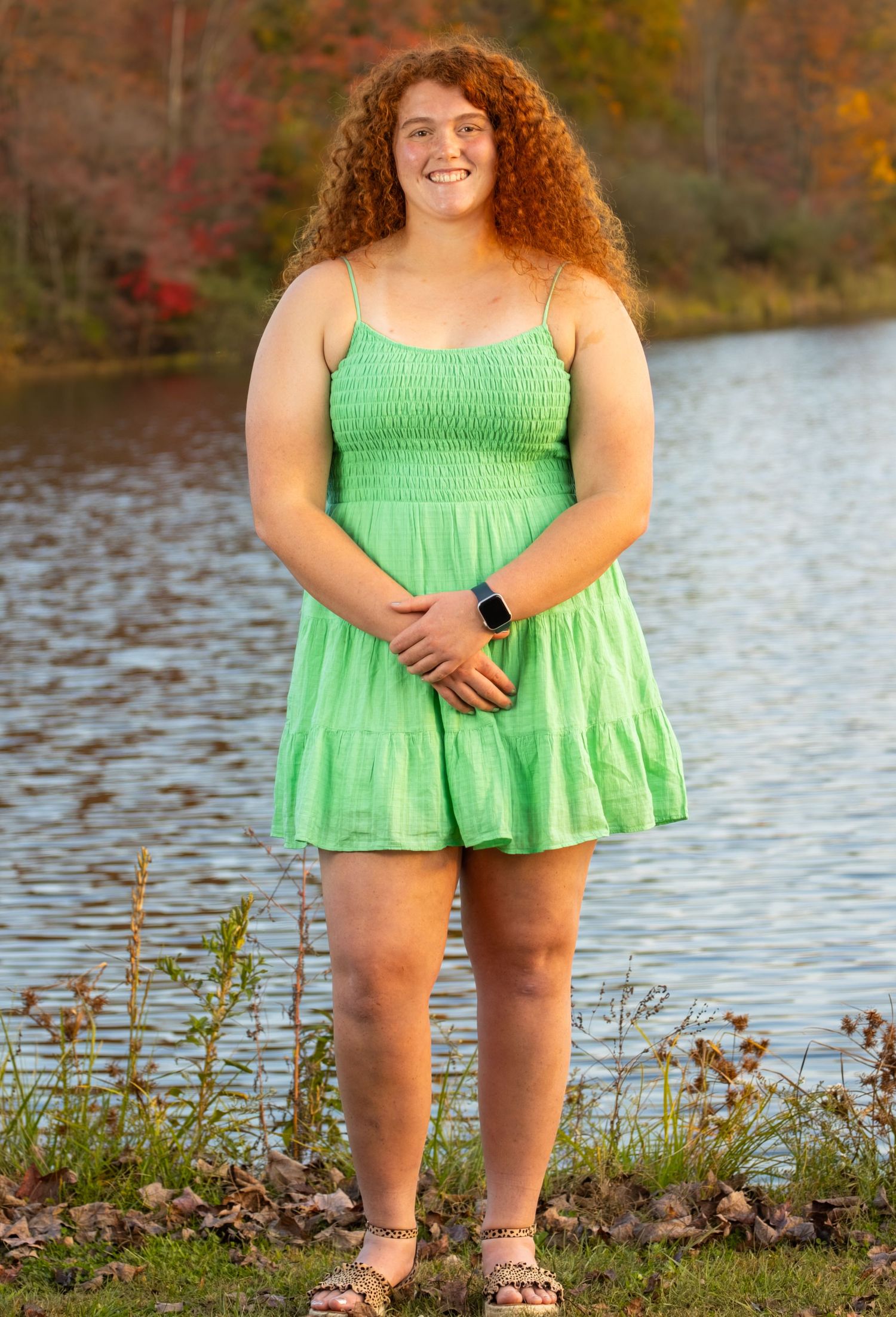 Teenage girl with curly red hair smiles in a green sundress standing beside a lake in autumn.