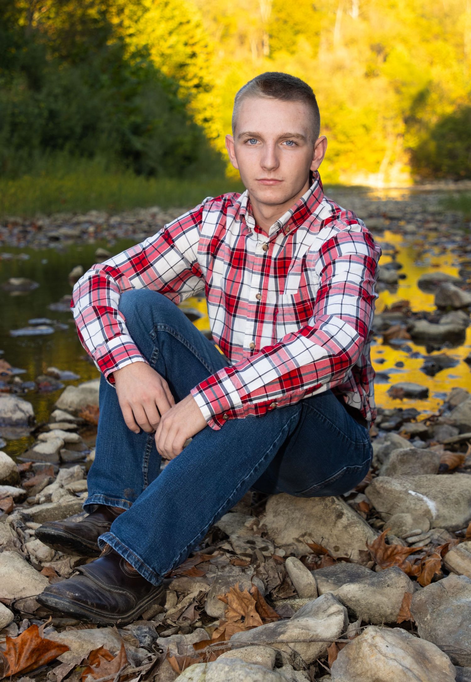 Young man in red plaid shirt and jeans sits on rocky riverbank surrounded by autumn foliage.