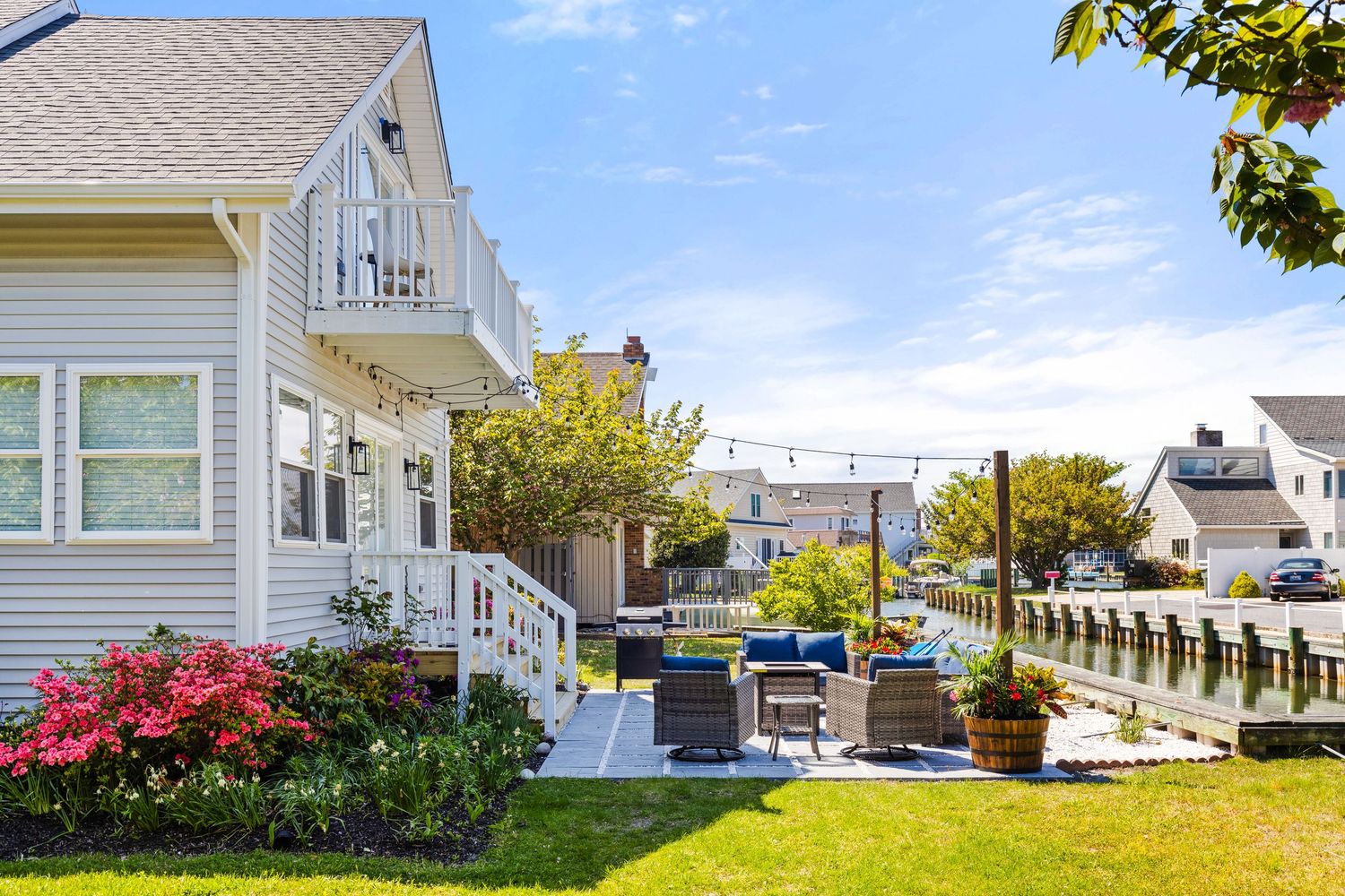 Charming coastal neighborhood with lush green lawn, colorful flower beds, and a sunny patio beside a scenic waterway.