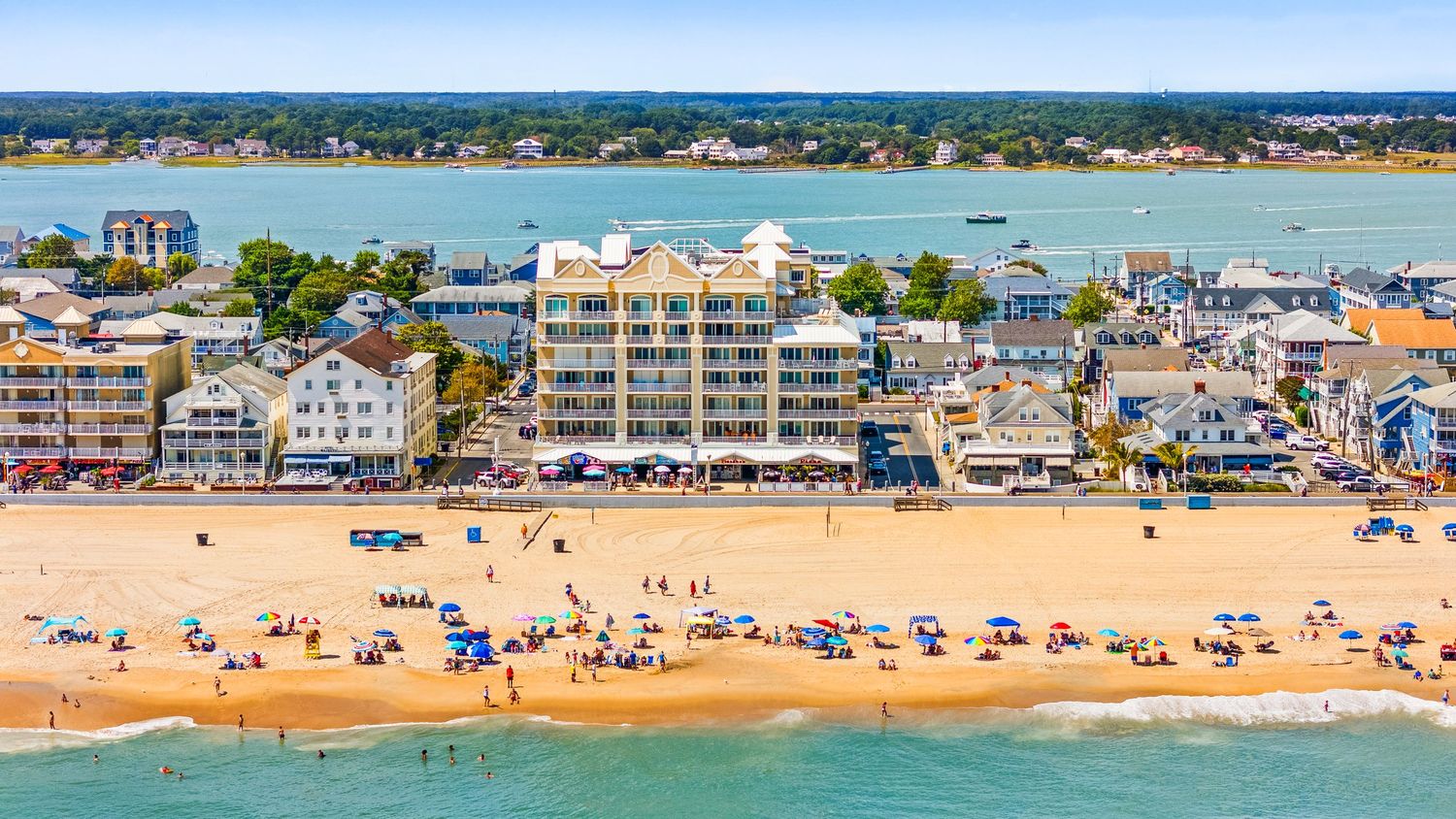 Aerial view of a busy beach town shoreline with colorful umbrellas, beachgoers, and multi-story coastal buildings.