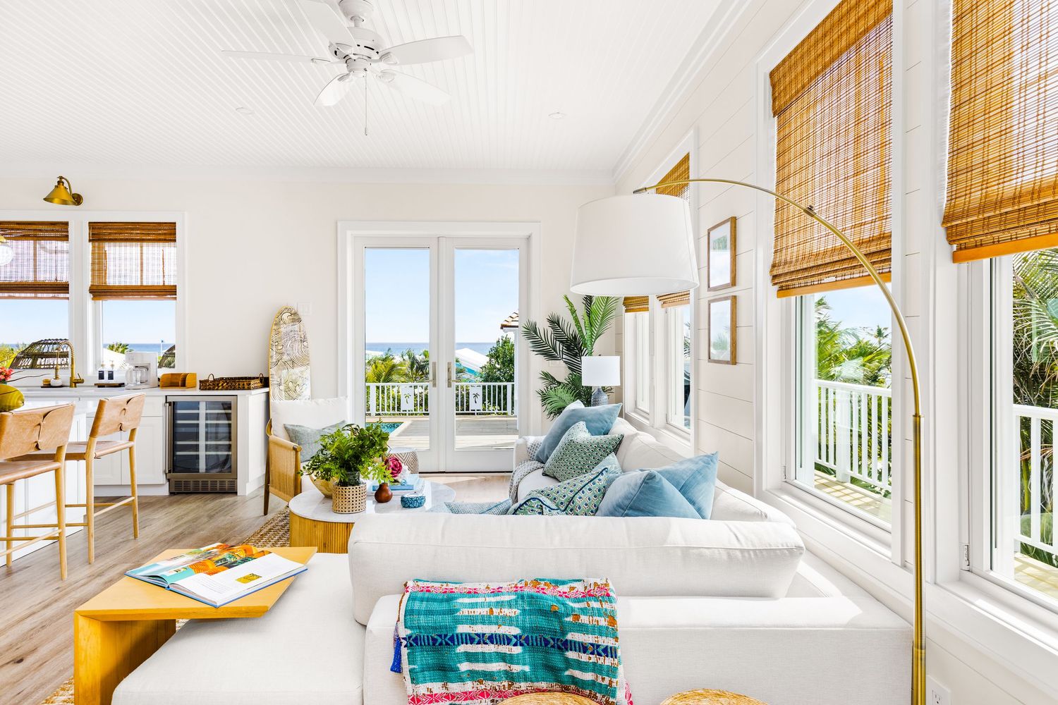 Bright coastal living room with white sofa, bamboo shades, blue accent pillows, hardwood floors, and a wine fridge in the background.