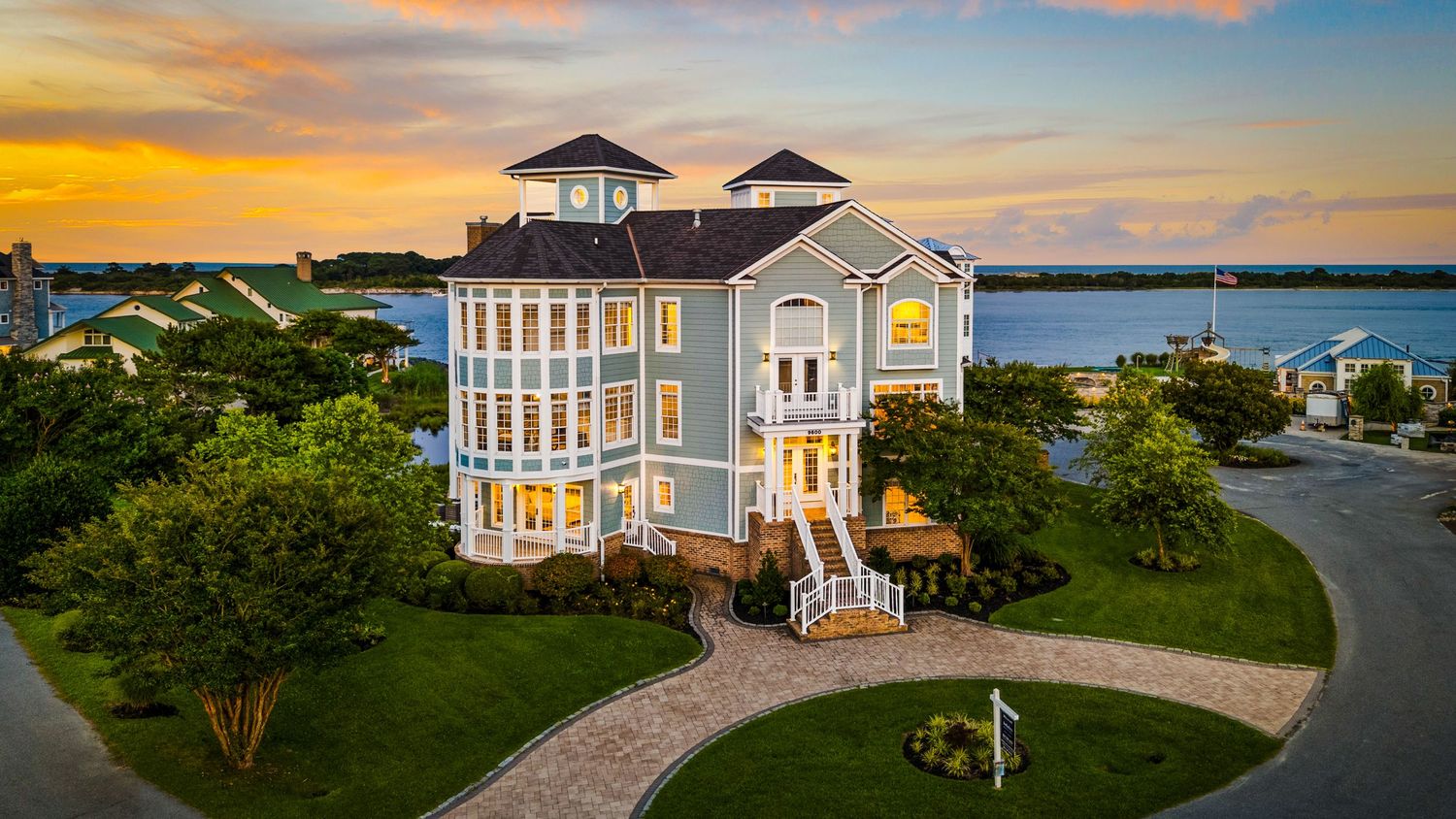 Stunning waterfront mansion with blue siding, wrap-around porches, and manicured lawn at sunset.