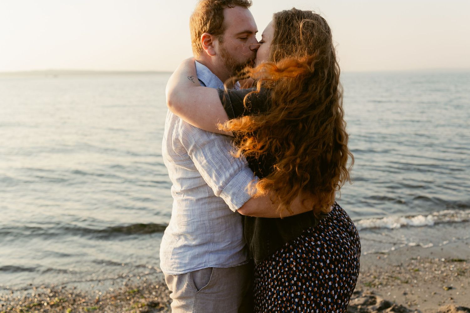Couple kissing and embracing by the water at Golden Gardens Beach in Seattle during golden hour.