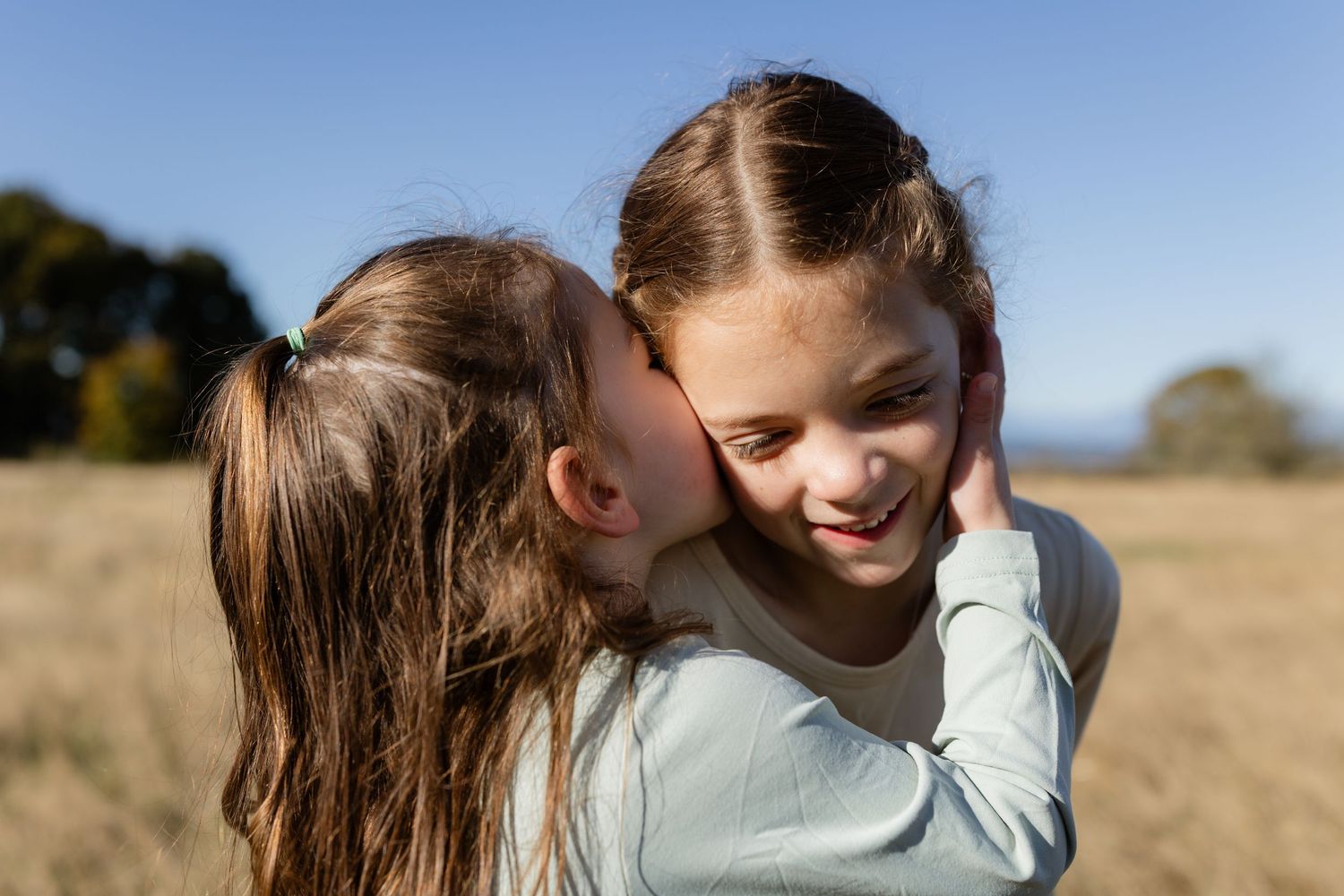 Two sisters sharing a sweet hug and a kiss on the cheek in a sunlit field at Discovery Park in Seattle.