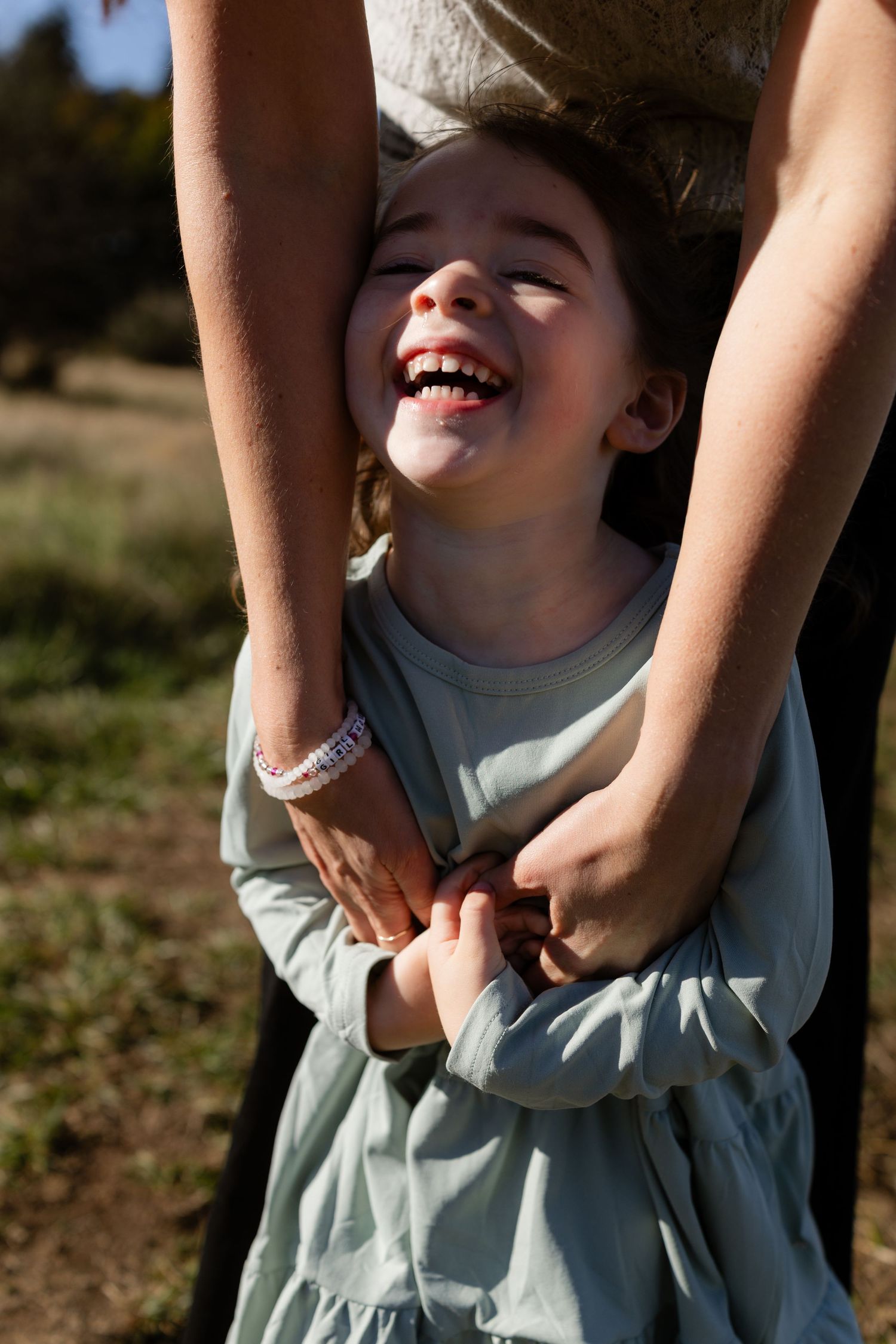 Young girl laughing as an adult holds her hands from behind during a playful family photoshoot at Discovery Park in Seattle.