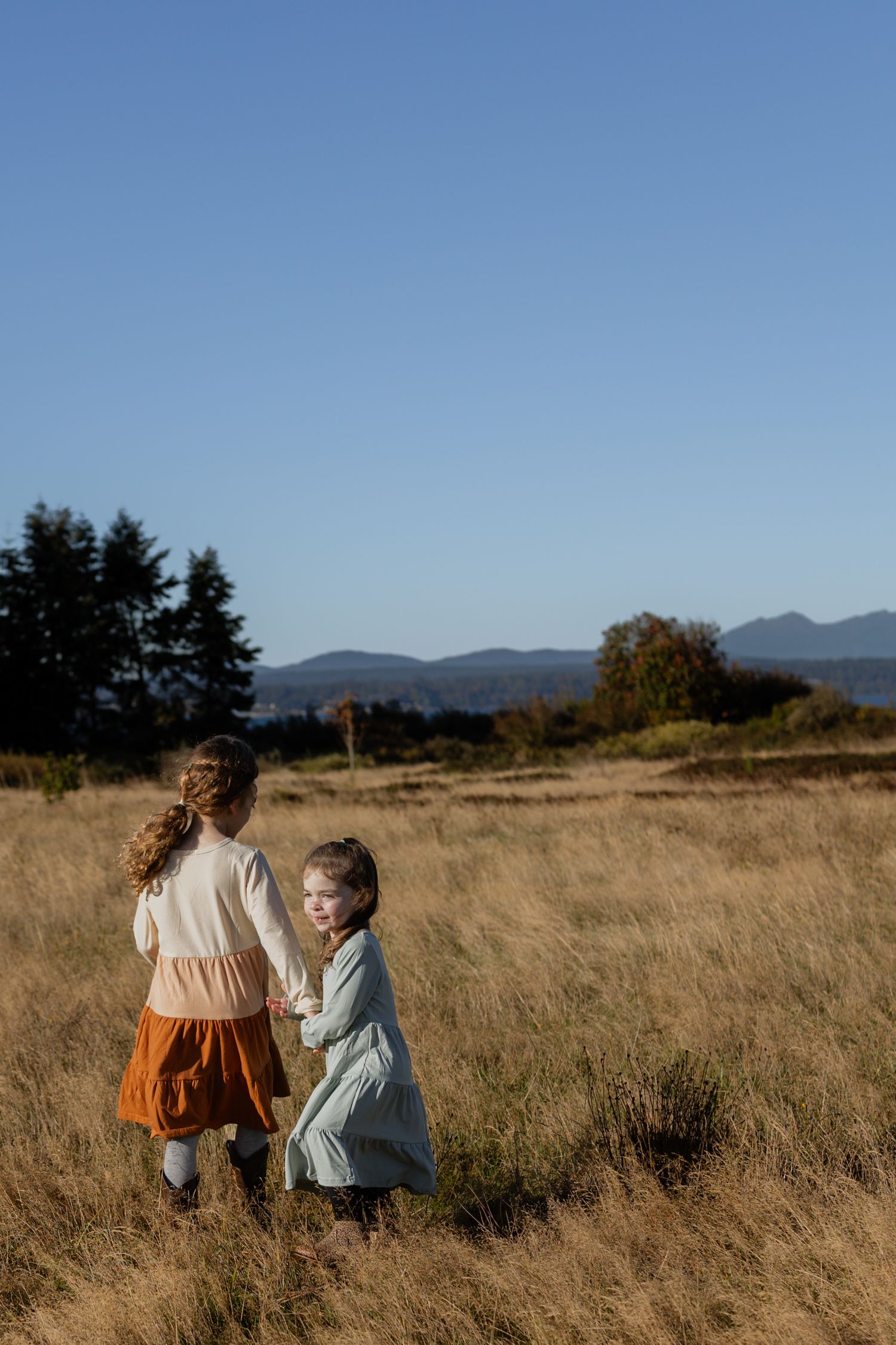 Two young sisters standing together in a wide, golden grassy field at Discovery Park in Seattle during a family photoshoot.