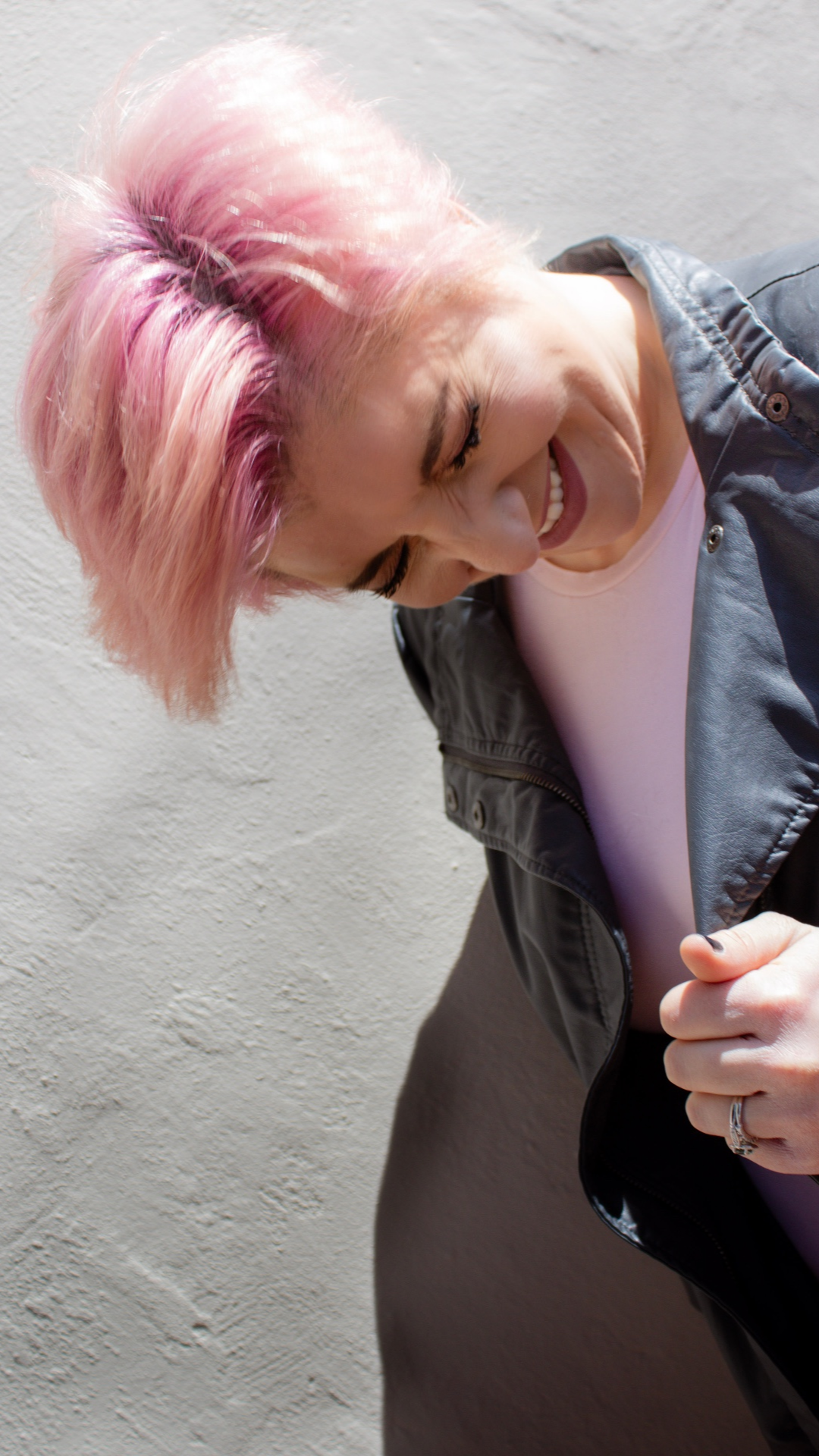 Creative portrait of a person with short pink hair leaning toward the camera against a neutral concrete background.