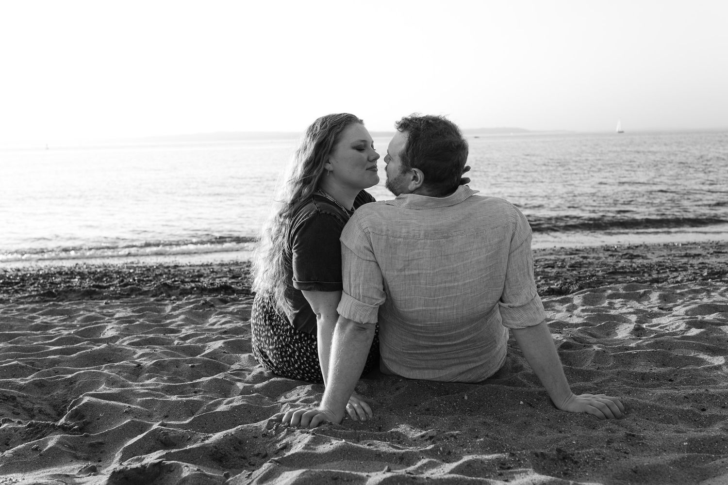Black-and-white portrait of a couple sitting close together on the sand at Golden Gardens Beach in Seattle, overlooking the water.