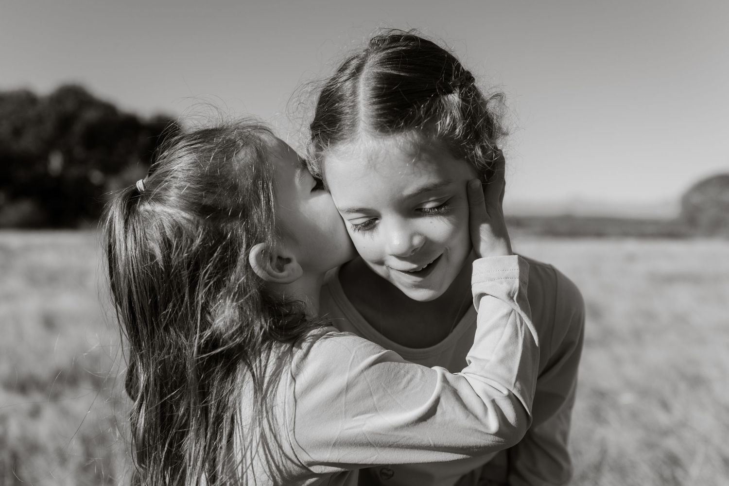 Black-and-white photo of two sisters sharing a kiss on the cheek in a grassy field at Discovery Park in Seattle.