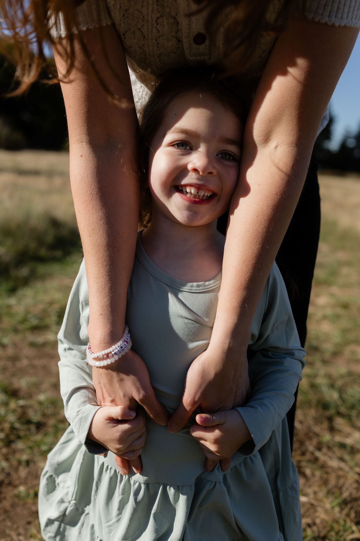 Young girl laughing as an adult holds her hands from behind during a candid family photography session at Discovery Park in Seattle.