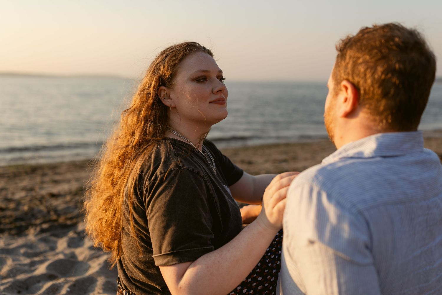 Candid couples portrait at Golden Gardens Beach in Seattle, with a woman looking at her partner beside the water at sunset.