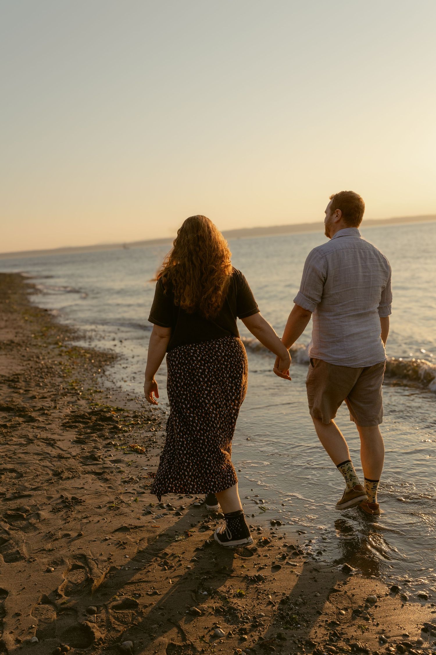Couple walking hand in hand along the rocky shoreline at Golden Gardens Beach in Seattle at sunset.